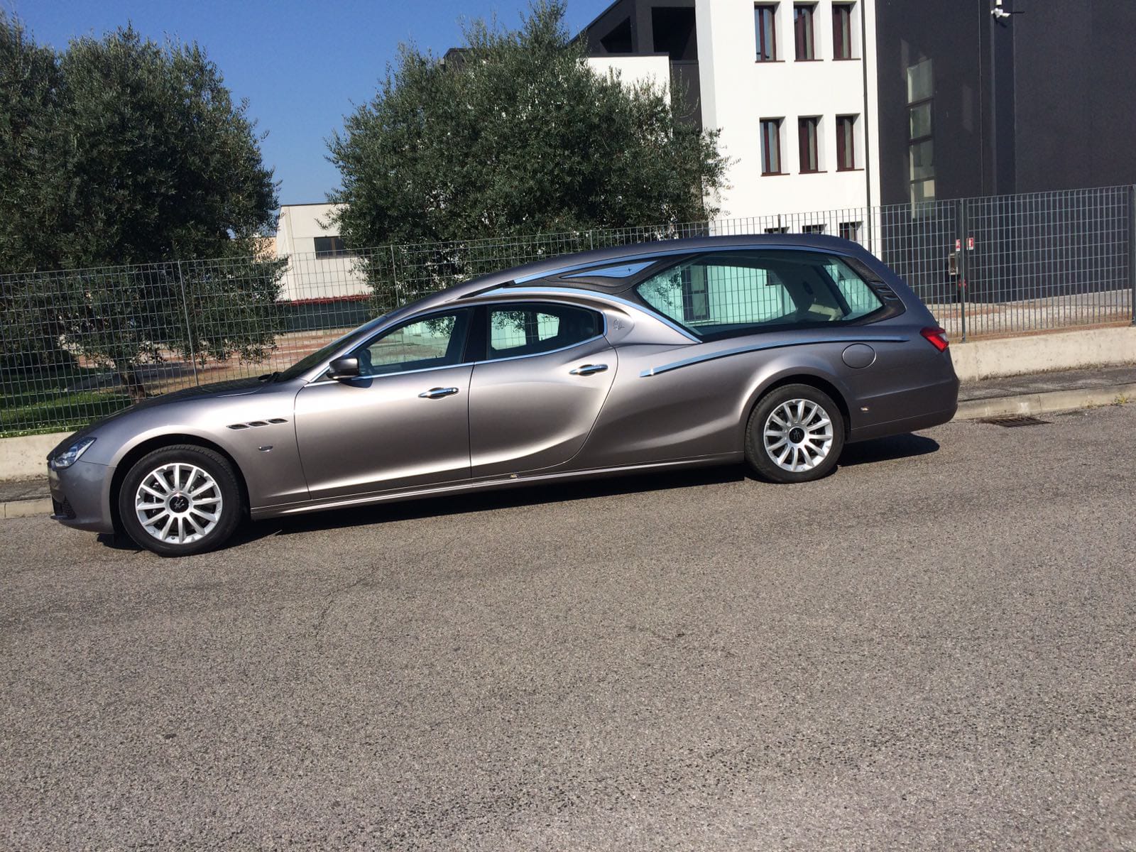 Carro funebre grigio parcheggiato su una strada asfaltata, con un edificio moderno e alberi sullo sfondo.