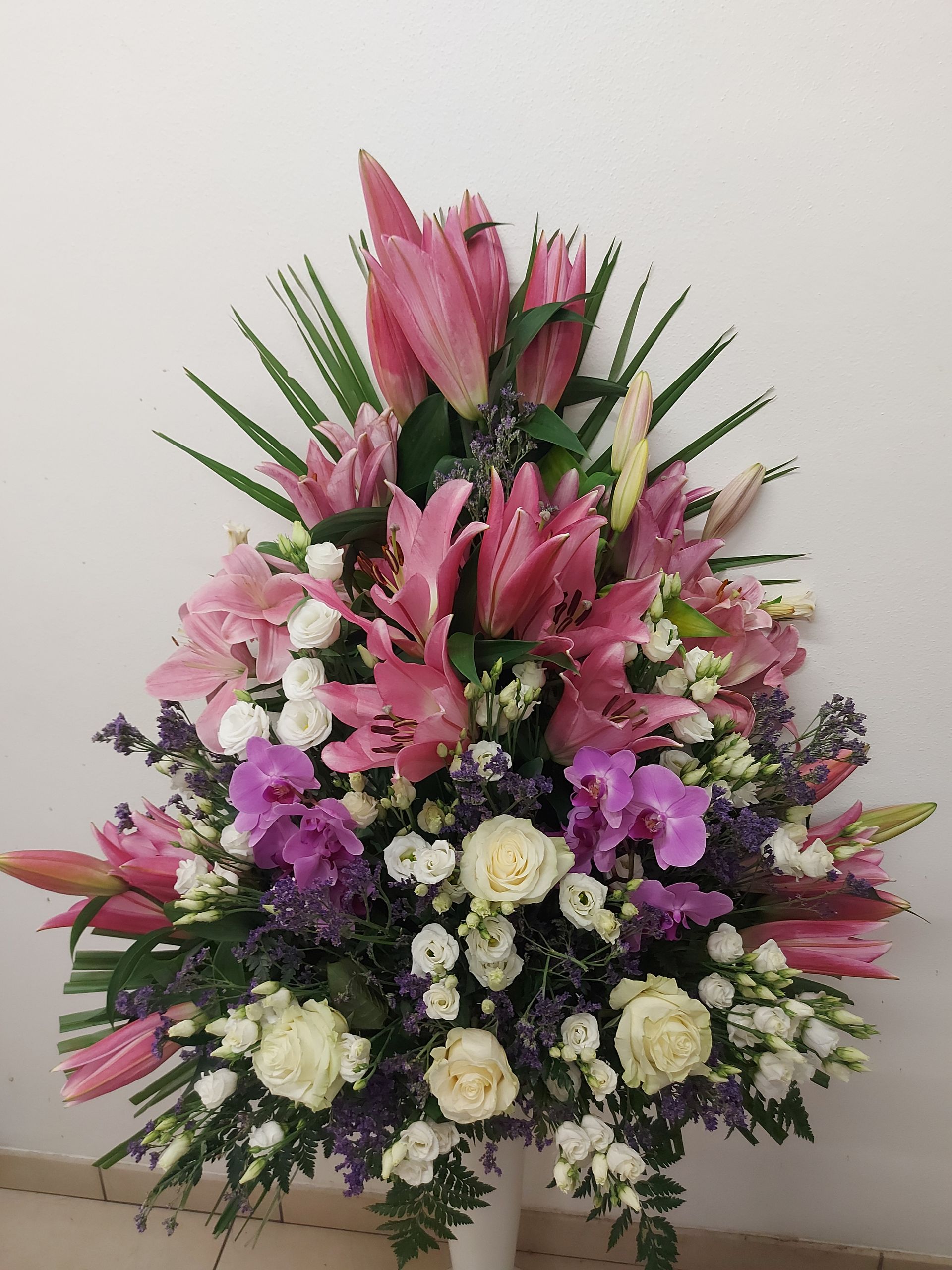 Composizione floreale: gigli rosa, rose bianche, orchidee viola, foglie verdi in un vaso bianco.