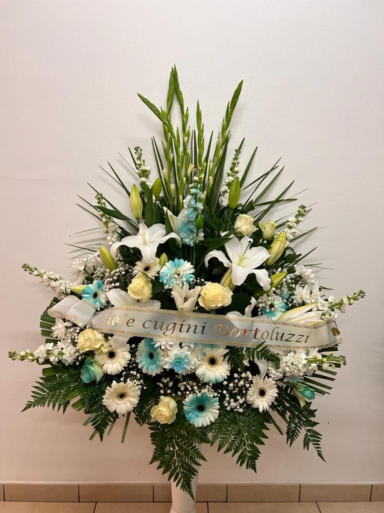 Composizione floreale in bianco, azzurro e verde; include gigli, margherite e nastro.
