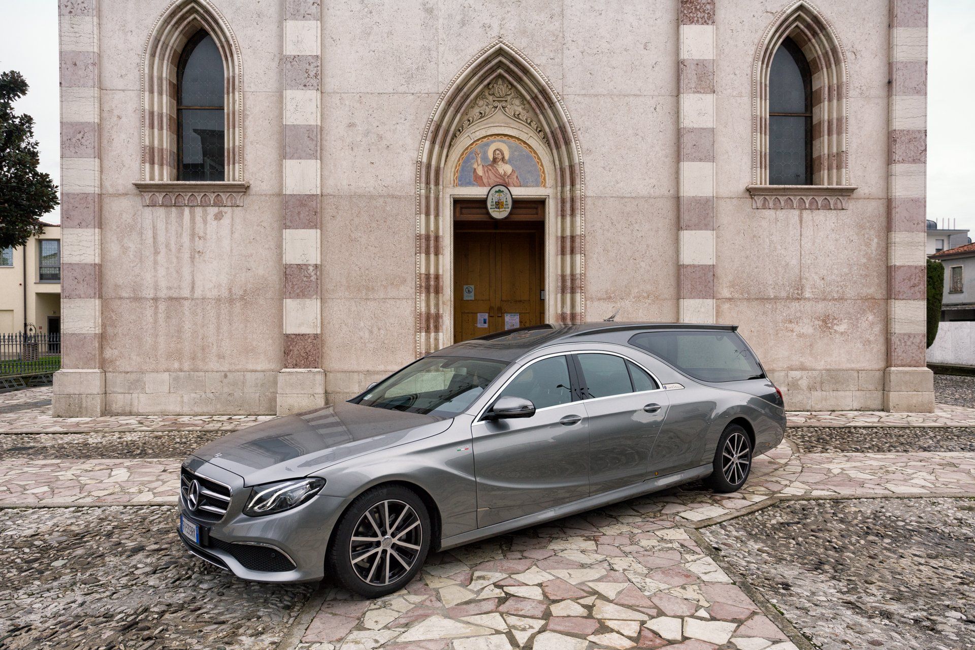 Un carro funebre argentato parcheggiato davanti a una chiesa di colore chiaro, con un portale ad arco decorativo e alte finestre.