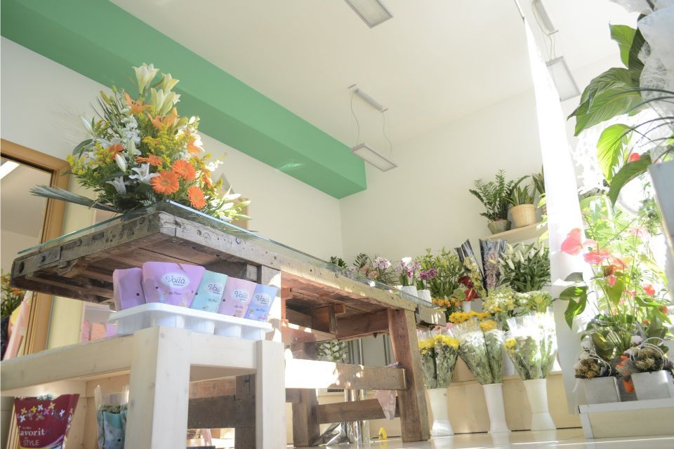 Interno di un negozio di fiori. Tavolo espositivo con composizioni floreali. Pareti bianche, accenti verdi e luce naturale intensa.