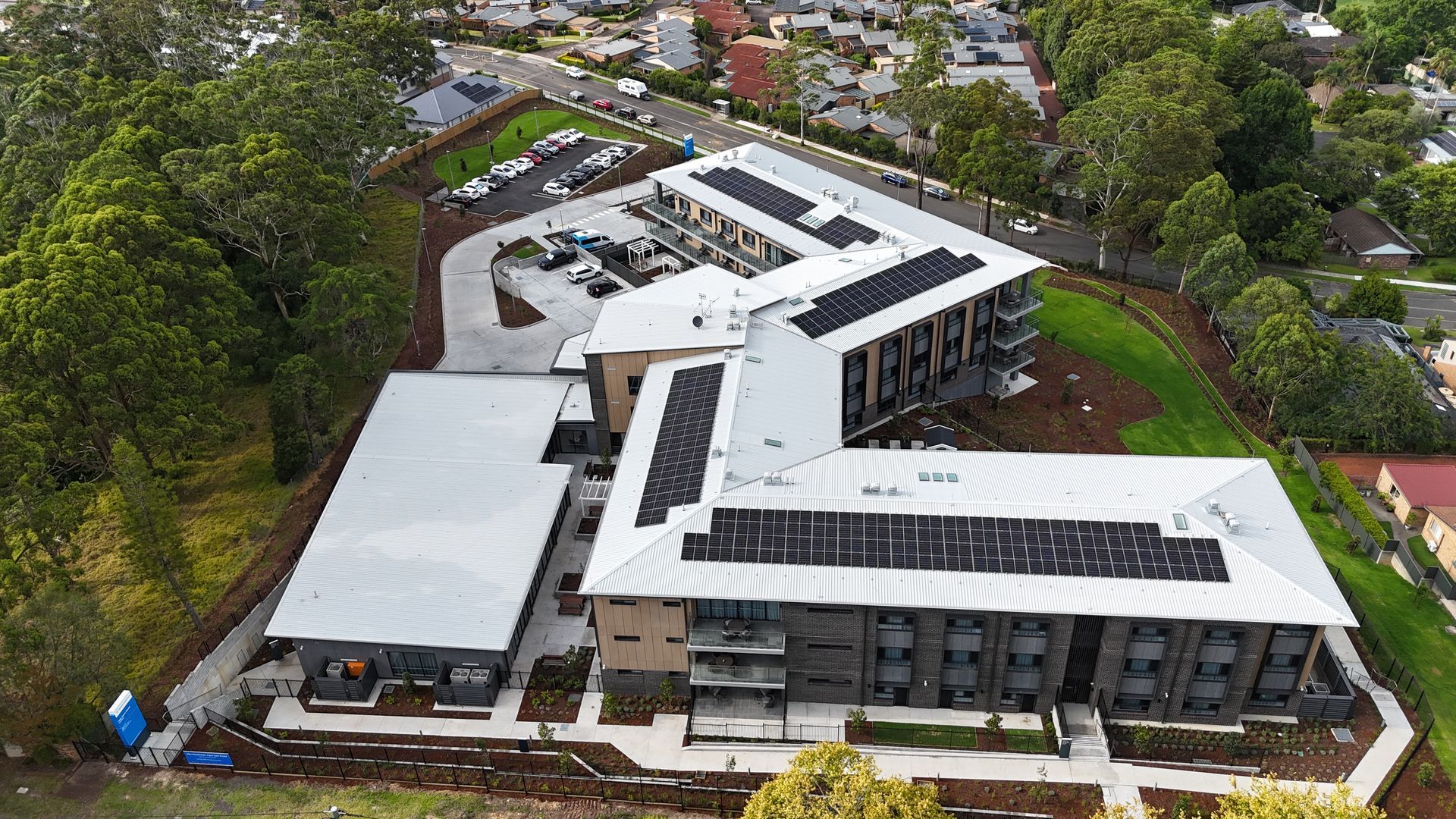 Aerial view of a modern building with solar panels on the roof and a surrounding parking lot and greenery.