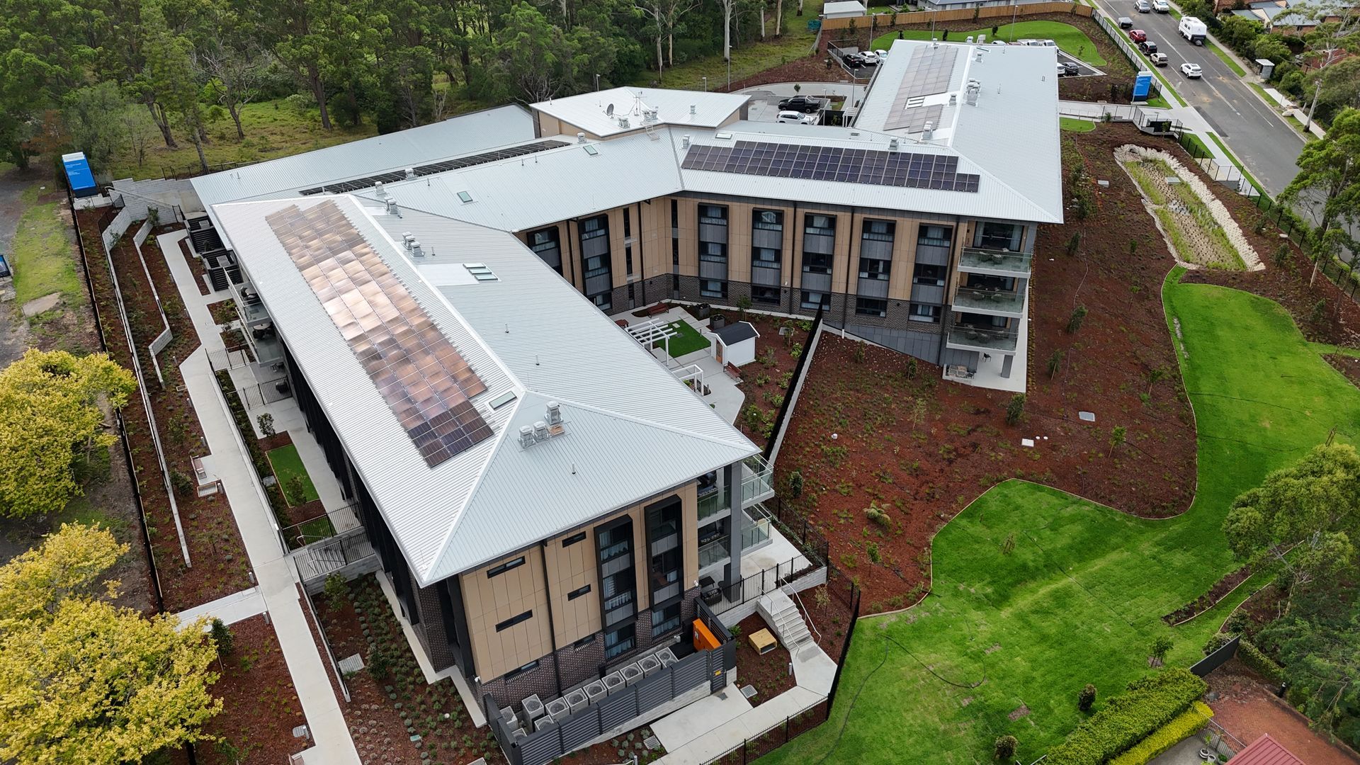 Aerial view of a modern multi-story building with a solar panel roof, next to a grassy hill and road.