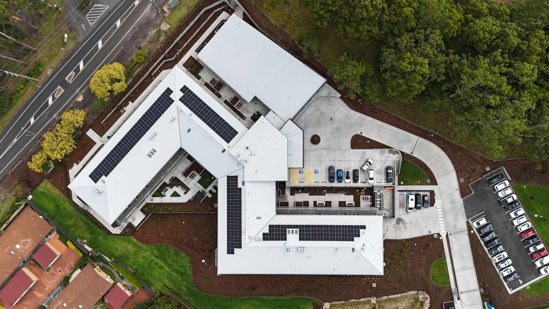 Aerial view of a modern building with solar panels on the roof, surrounded by landscaping, a road, and a parking lot.