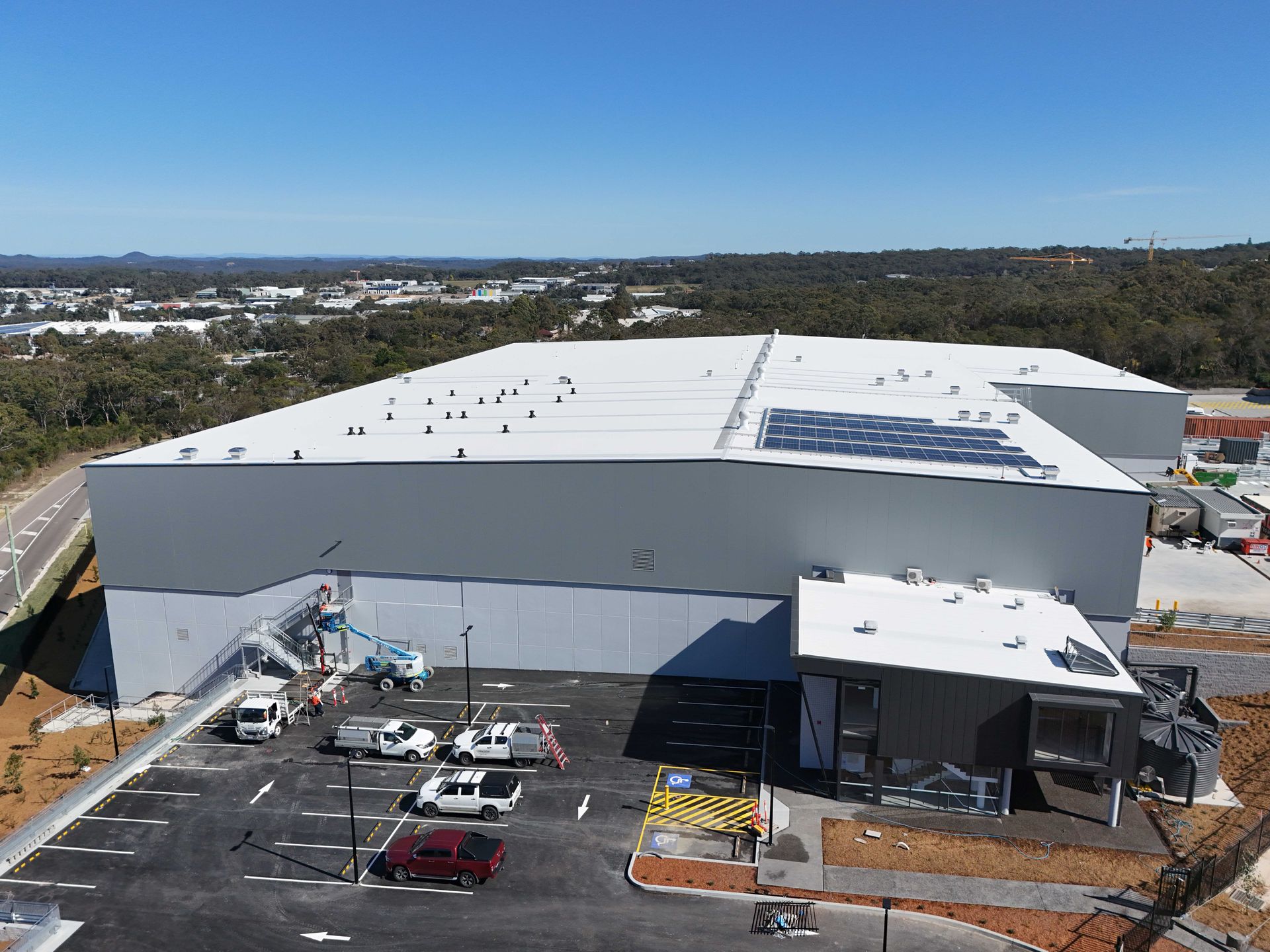 Large gray industrial building with solar panels, parked vehicles, and surrounding greenery.