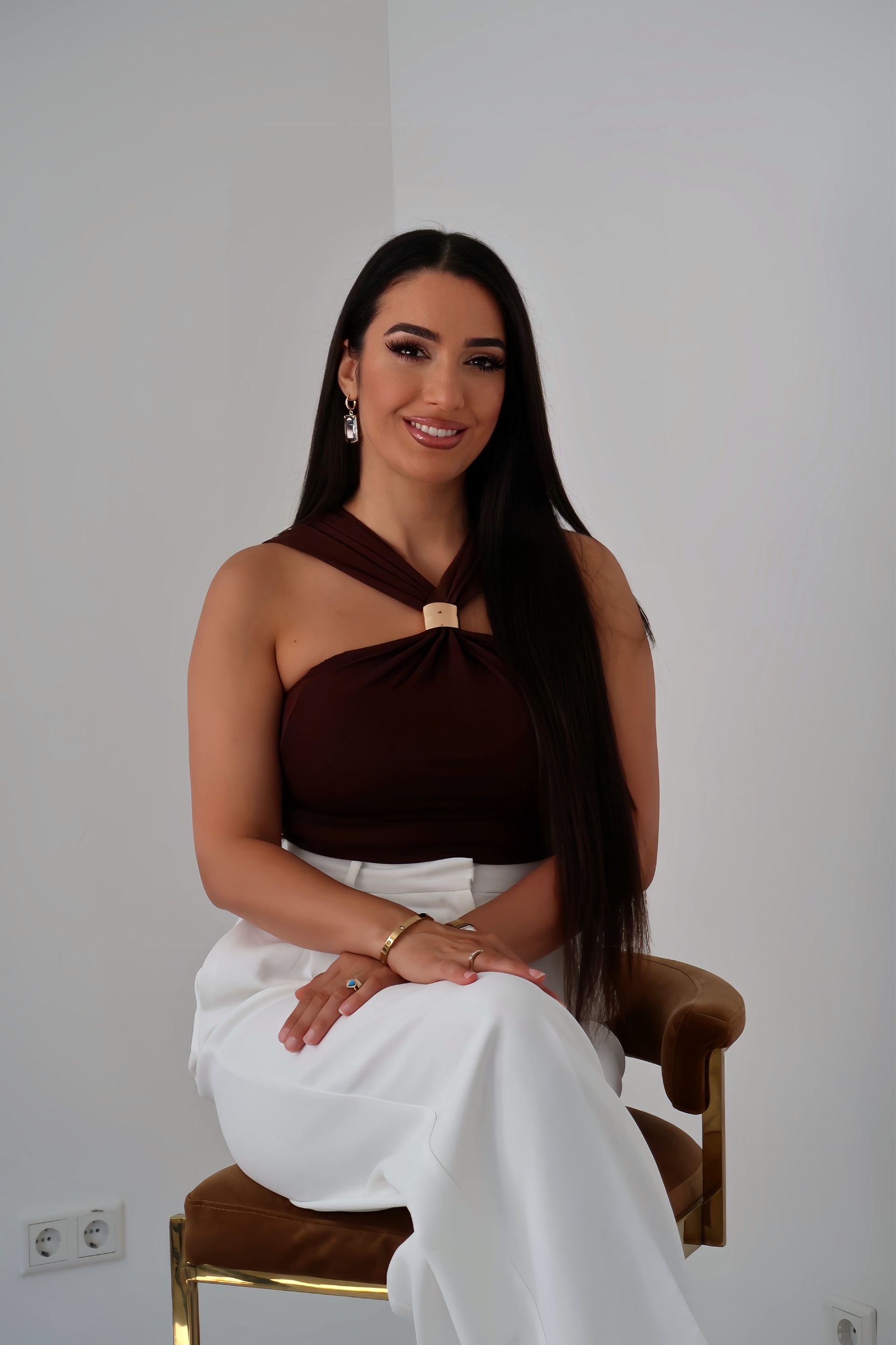 Frau mit langem dunklem Haar, lächelnd, mit braunem Oberteil und weißen Hosen bekleidet, sitzt auf einem goldenen Stuhl.