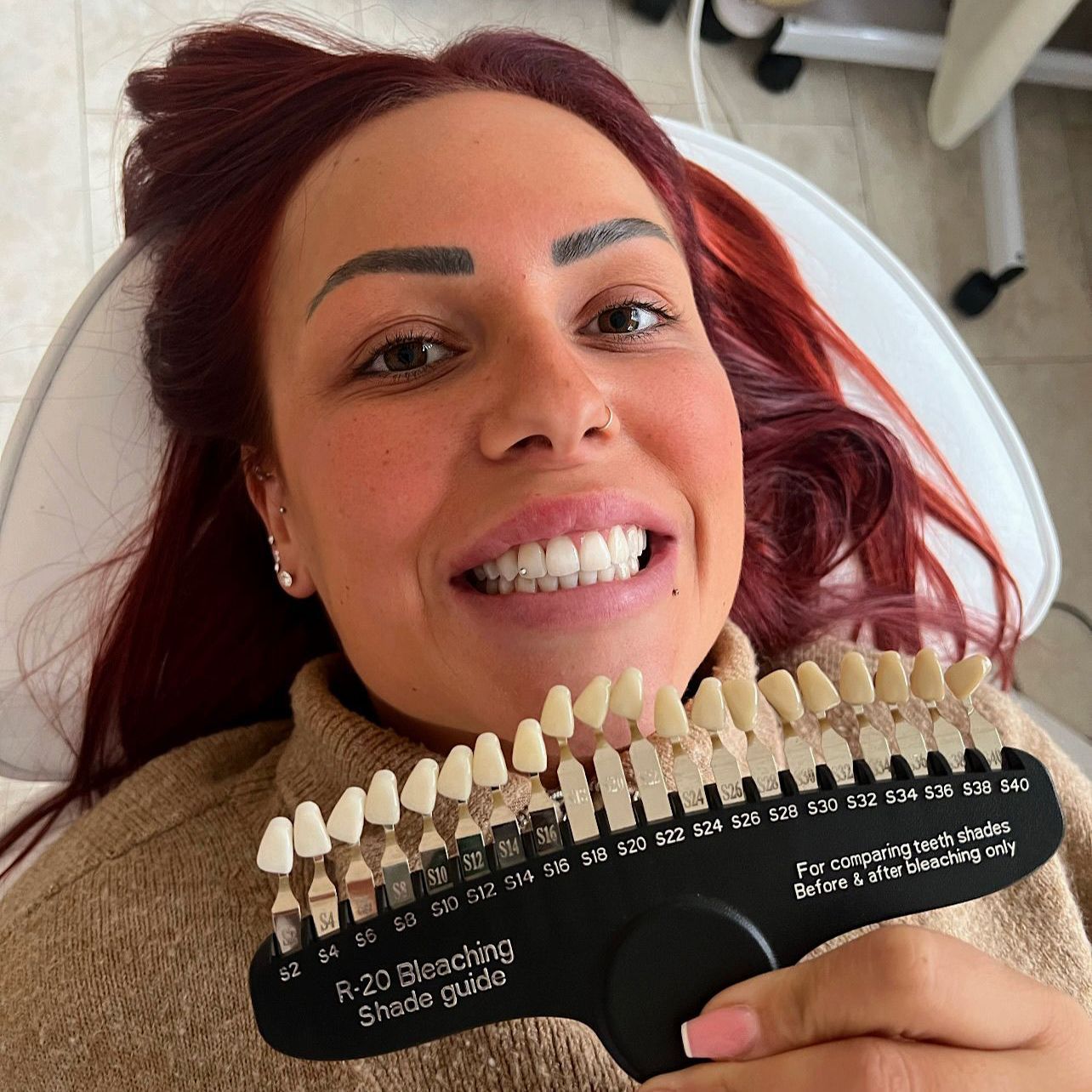 Lächelnde Frau mit roten Haaren, die in einer Zahnarztpraxis eine Zahnfarbskala hält.