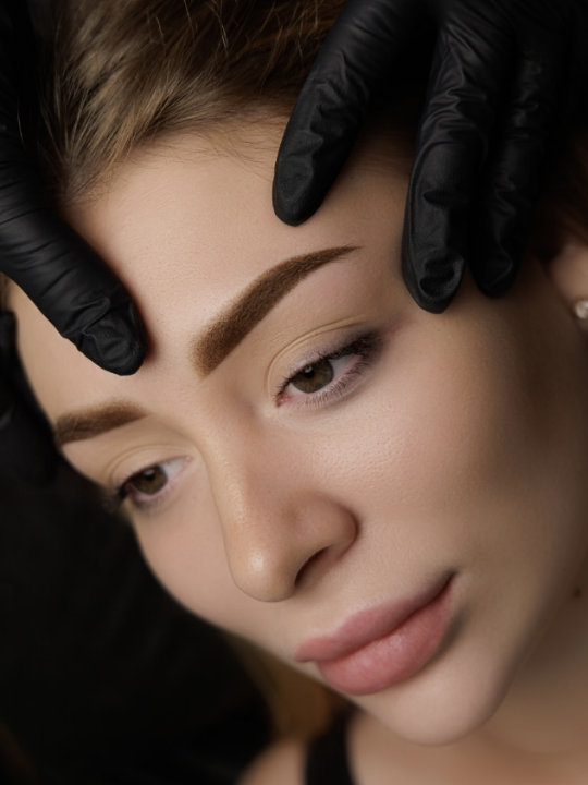 Einer Frau werden von einer Person mit schwarzen Handschuhen die Augenbrauen zurechtgemacht.