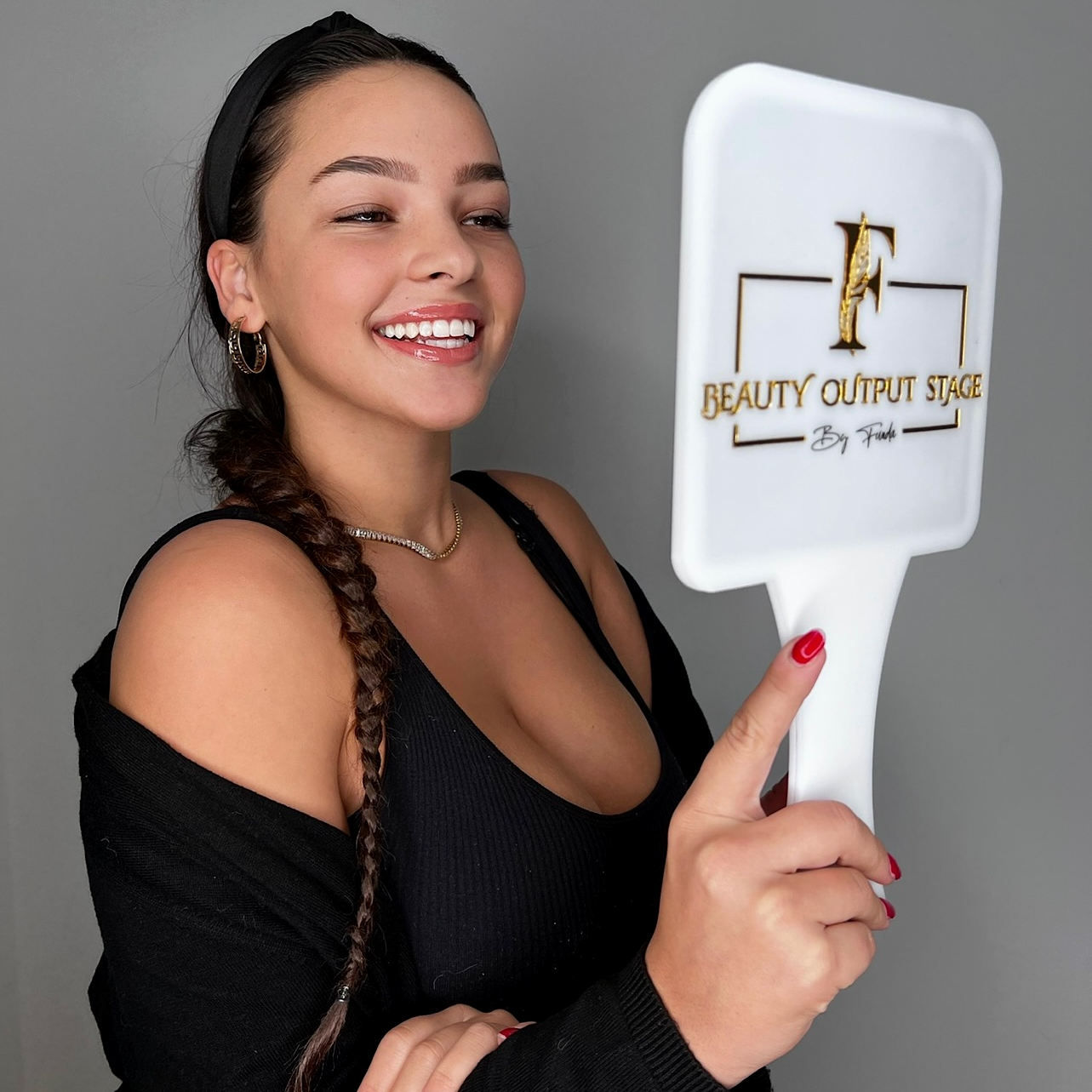 Frau lächelt, während sie einen weißen Handspiegel mit einem Logo hält und ein schwarzes Outfit trägt.