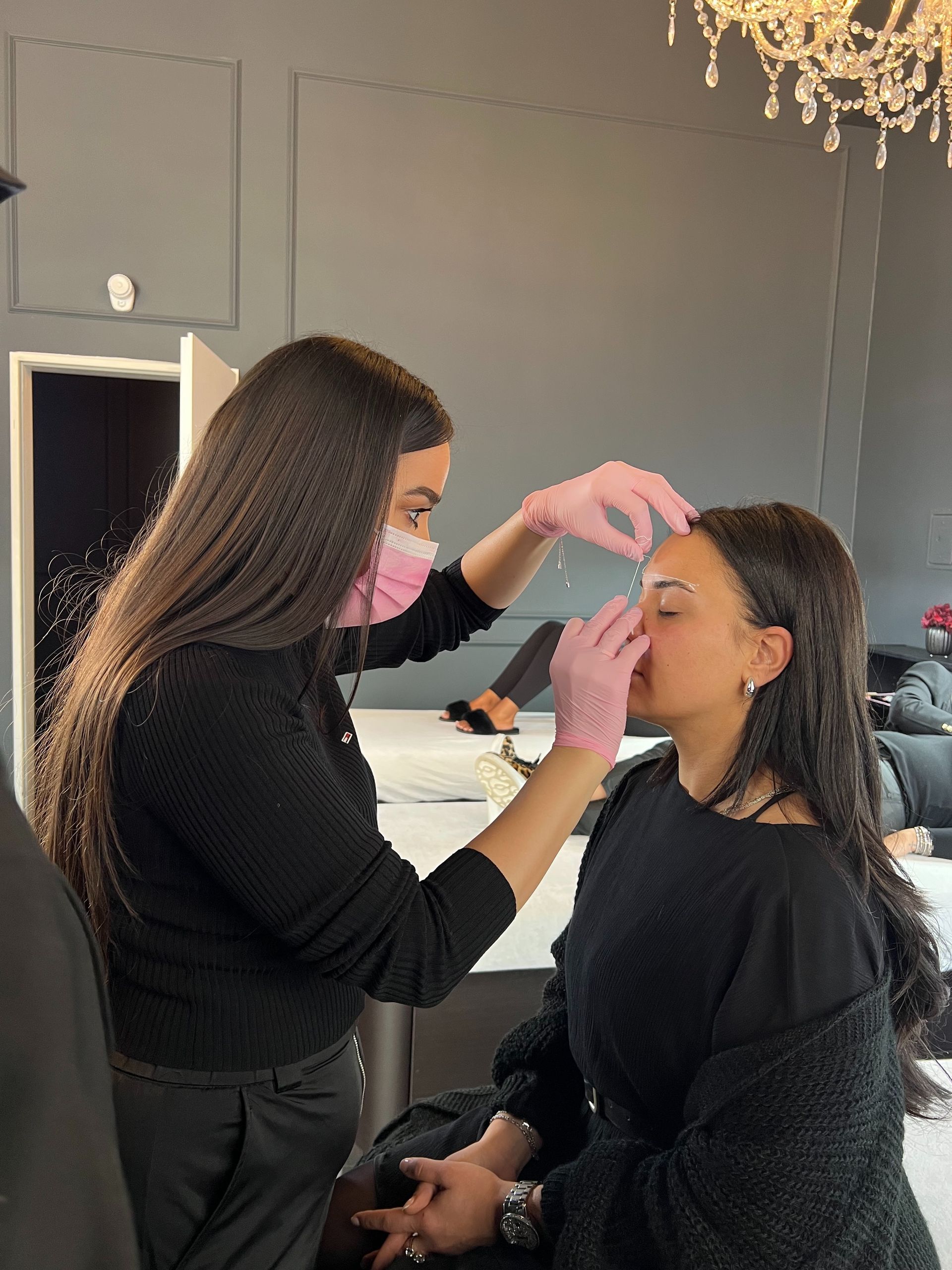 Eine Frau mit rosa Handschuhen untersucht in einem Salon das Gesicht einer anderen Frau. Beide tragen schwarz-rosa Masken.