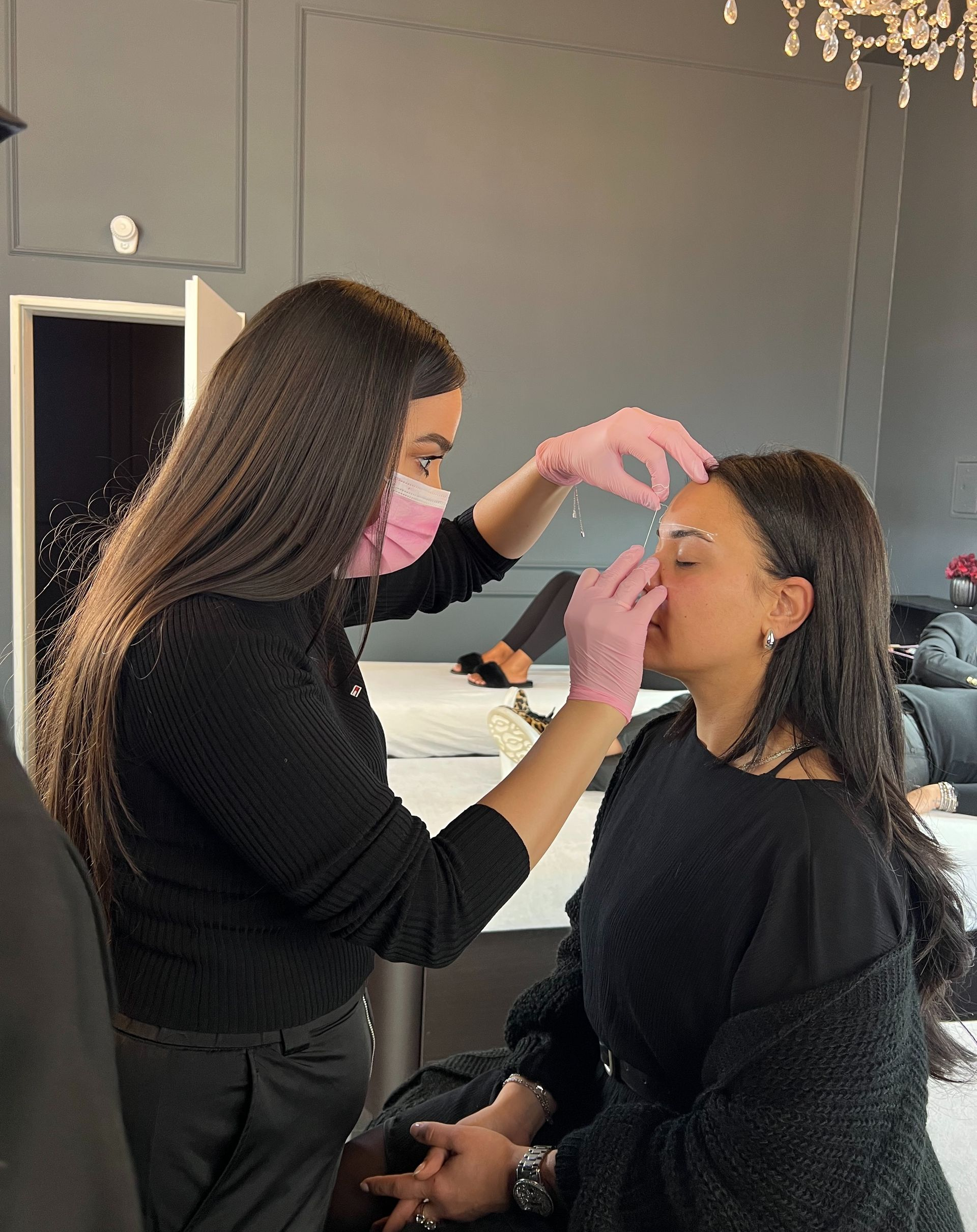 Frau mit rosa Handschuhen zupft einer Kundin in einem Salon die Augenbrauen zu. Beide tragen Masken, der Raum ist grau.