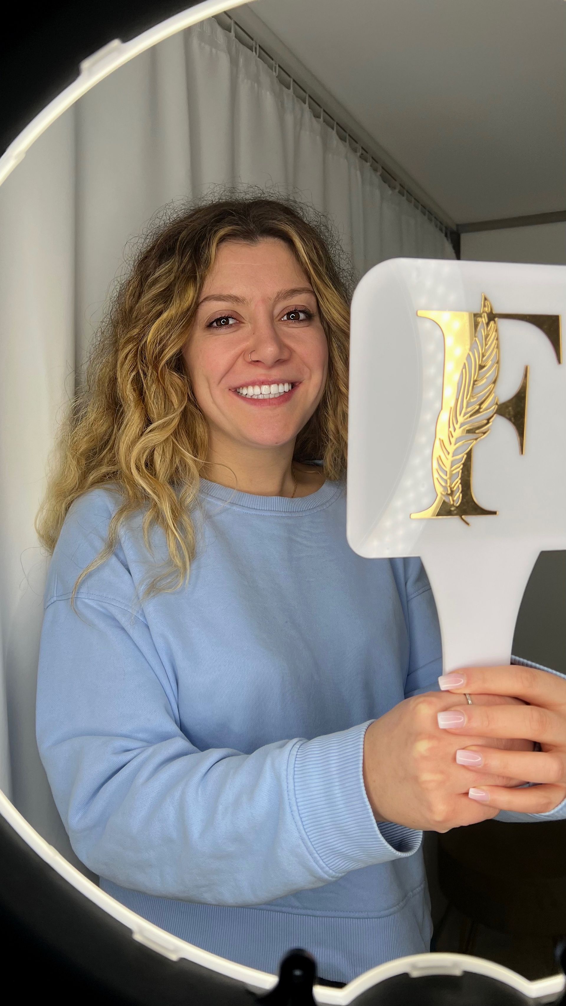 Frau im blauen Pullover lächelt und hält einen Handspiegel mit einem goldenen „F“ in einem hell erleuchteten Raum.