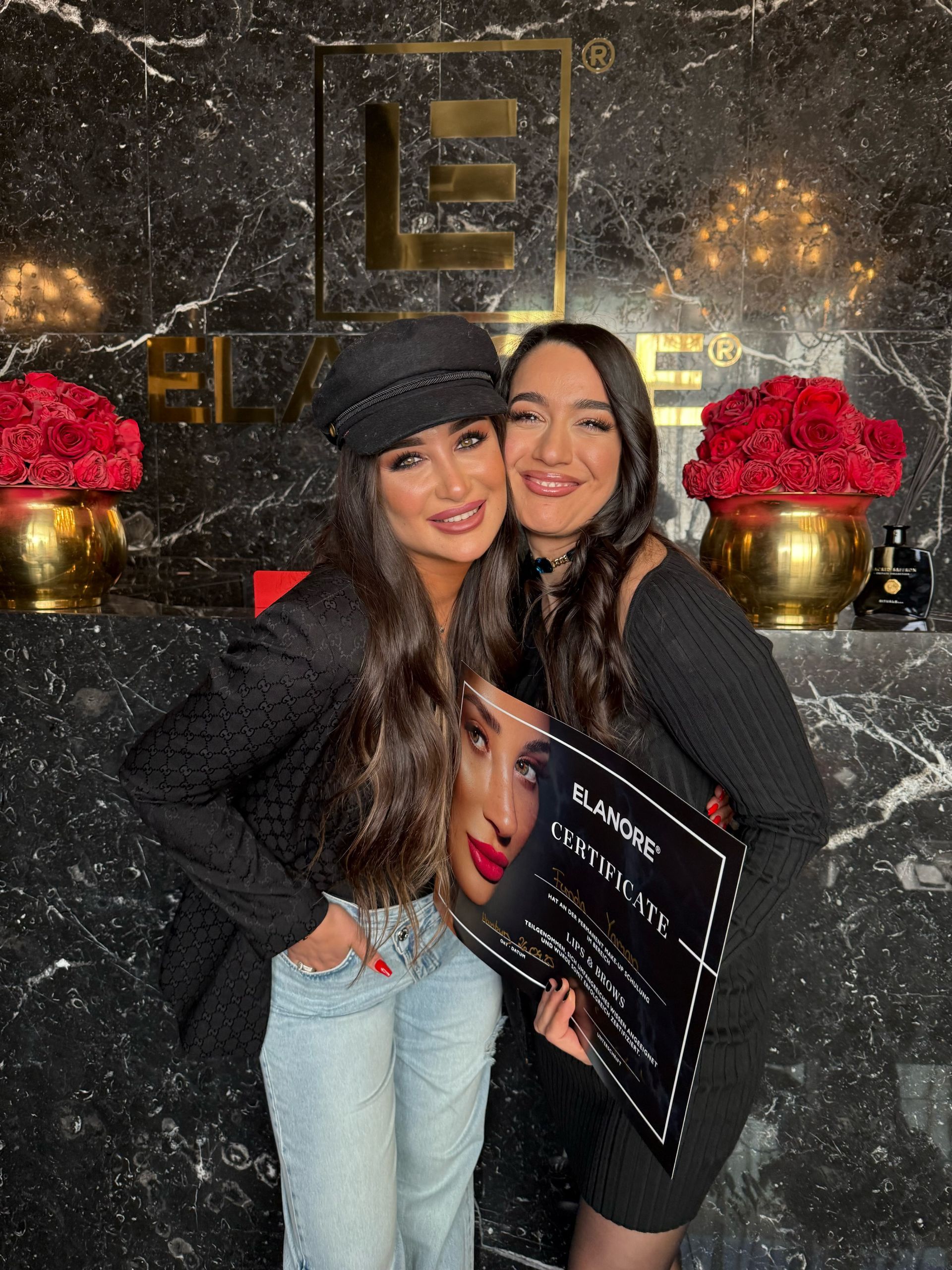 Zwei Frauen posieren vor einem Logo mit einer Urkunde, die eine trägt einen schwarzen Hut und lächelt, die andere lächelt.