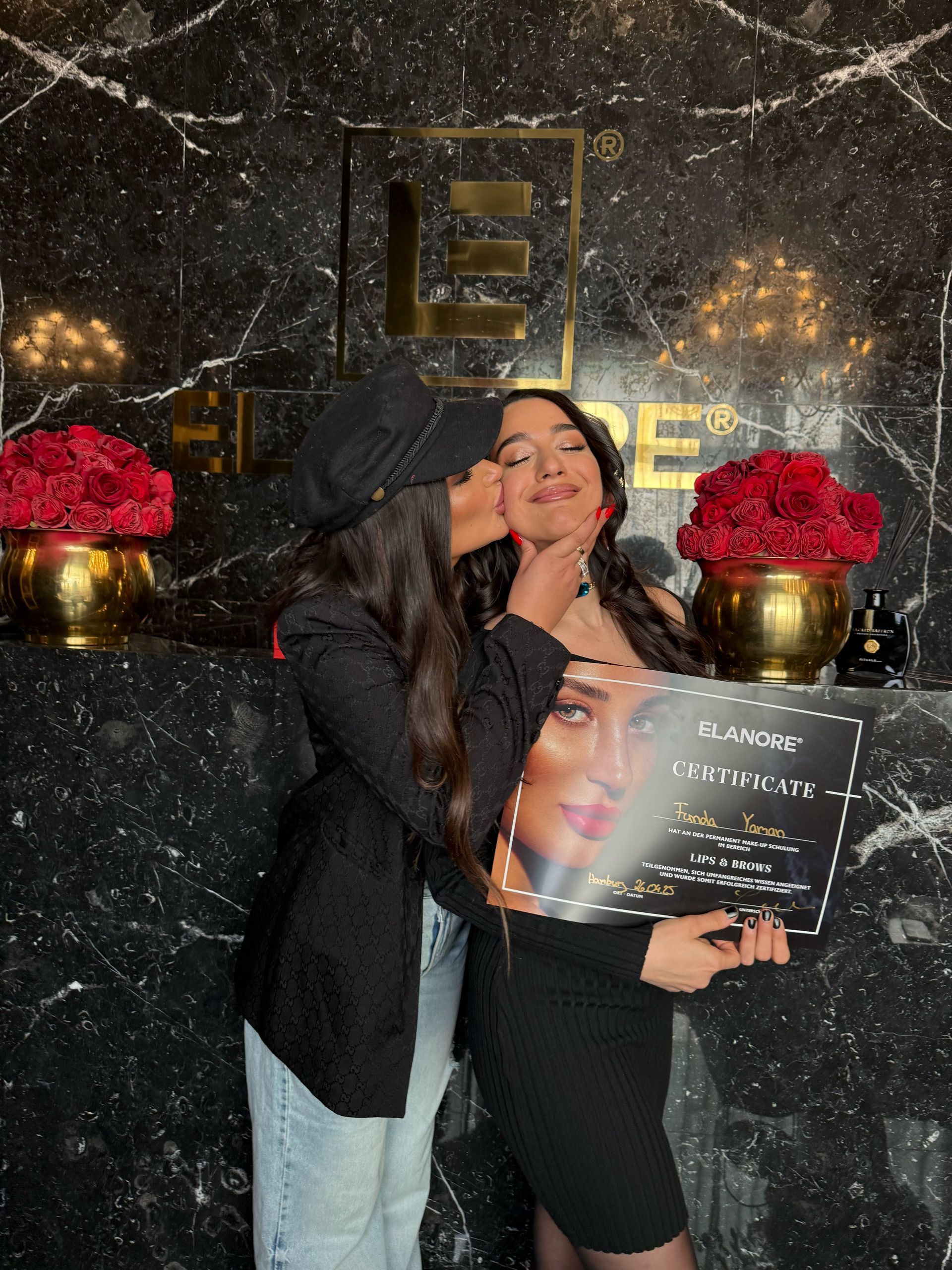 Zwei Frauen feiern. Eine küsst die andere und hält eine Urkunde vor einem floralen Hintergrund.