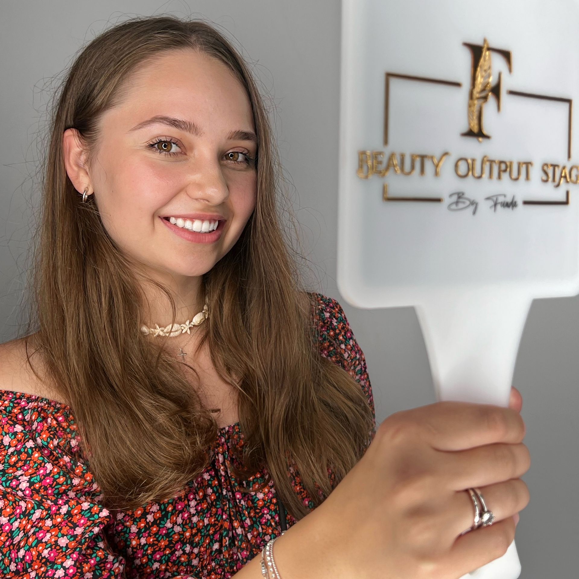 Junge Frau mit geblümtem Hemd lächelt und hält einen weißen Spiegel mit dem Logo „Beauty Output Stage“.