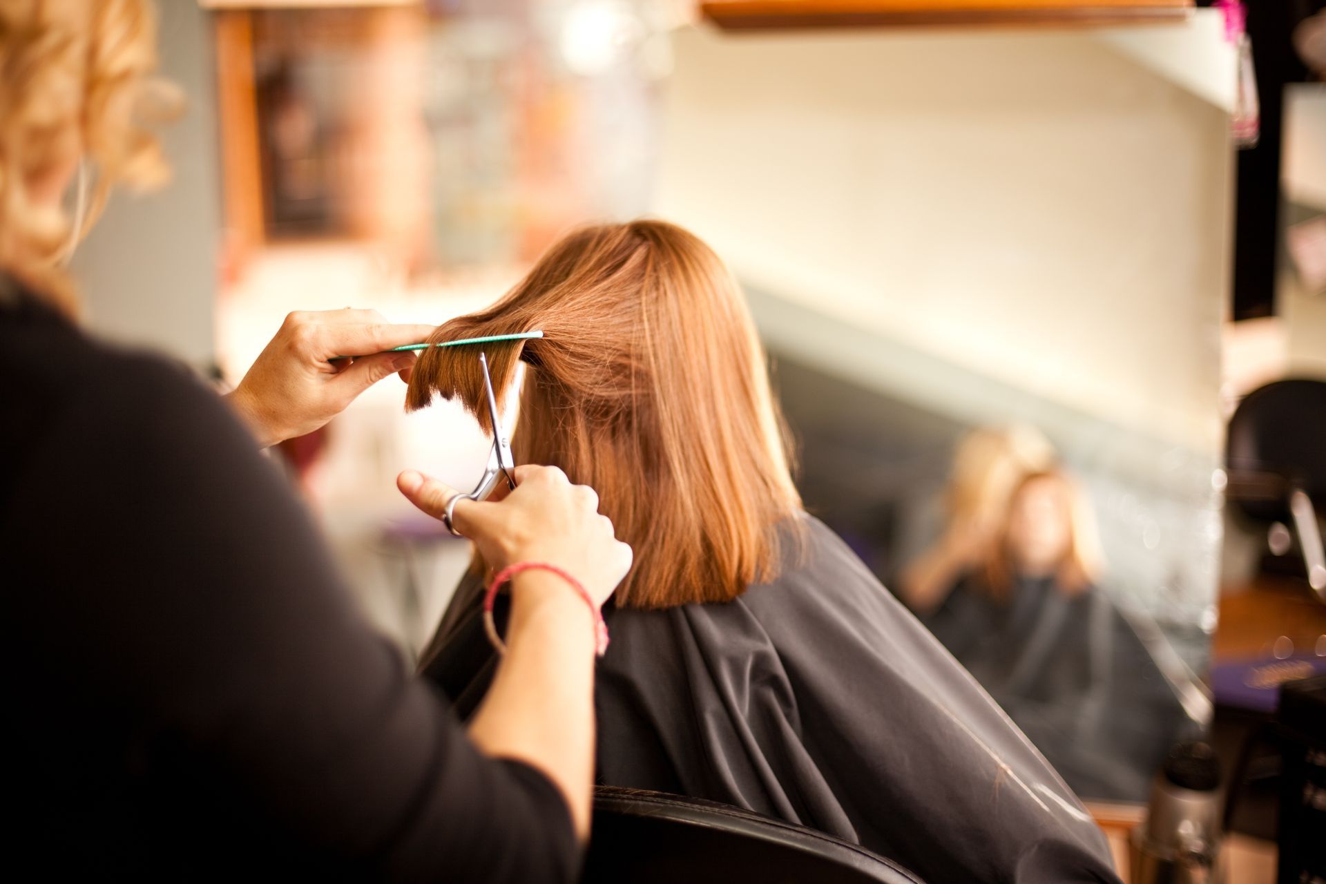 Hairdresser cutting a client's red hair with scissors in a salon, reflected in the mirror.