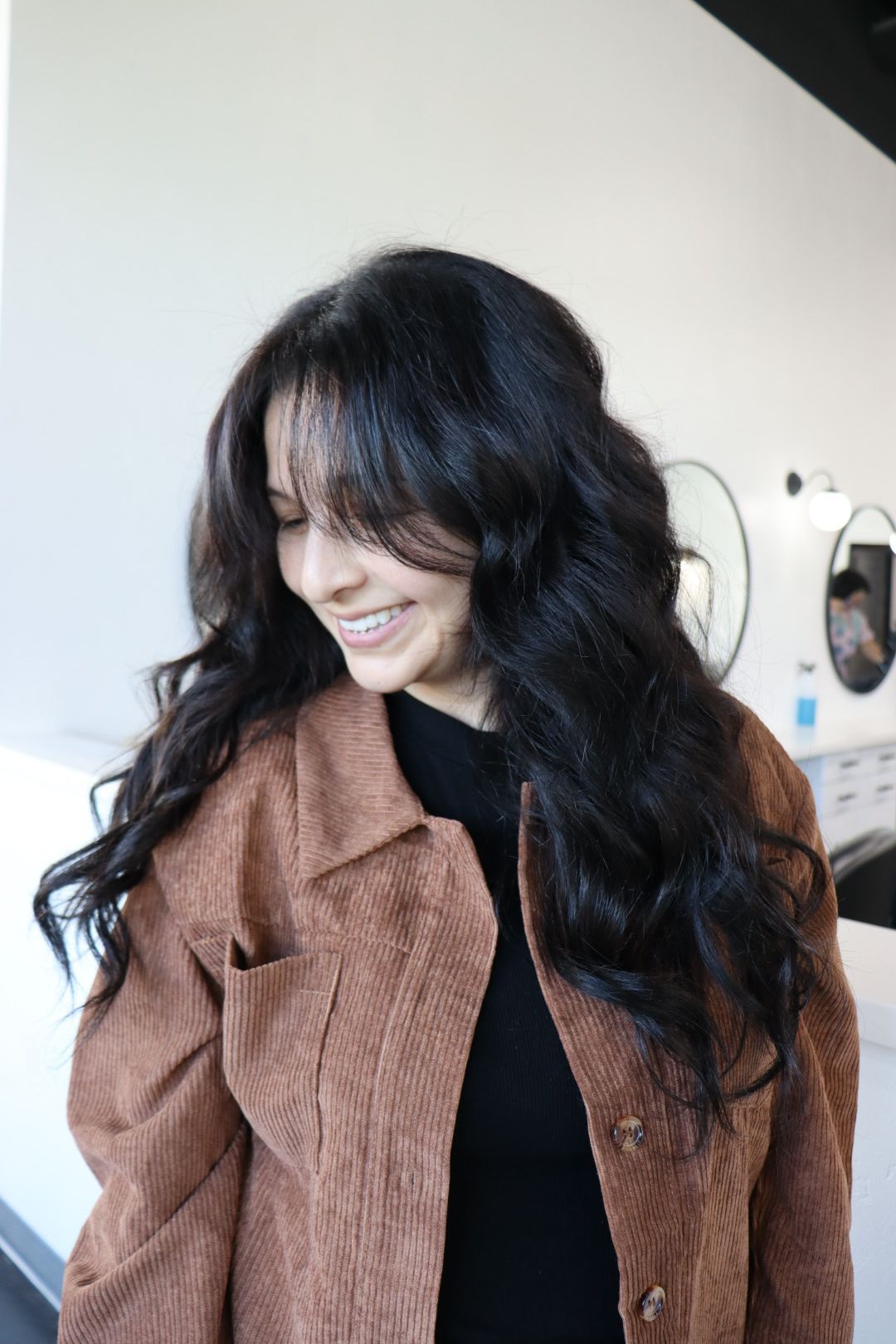 A woman with long black hair is wearing a brown jacket and smiling.