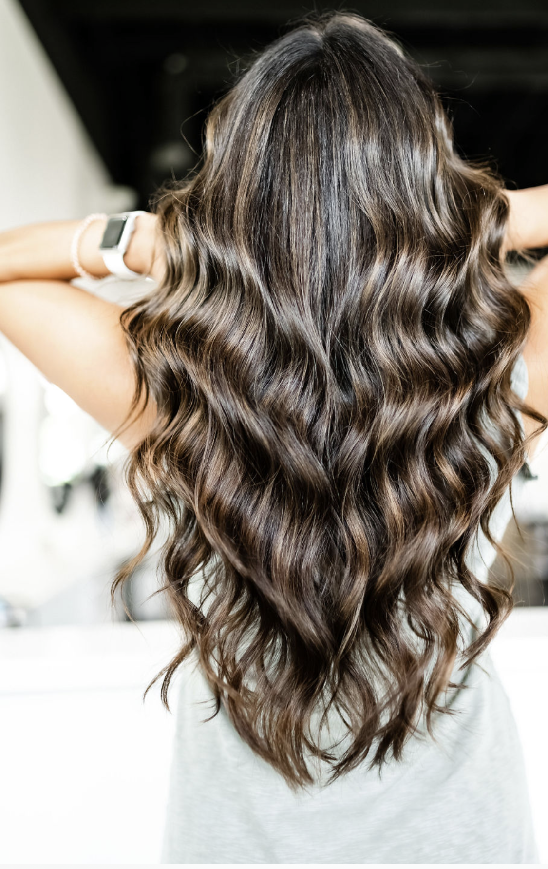 A woman with long wavy hair is standing with her hands behind her head.