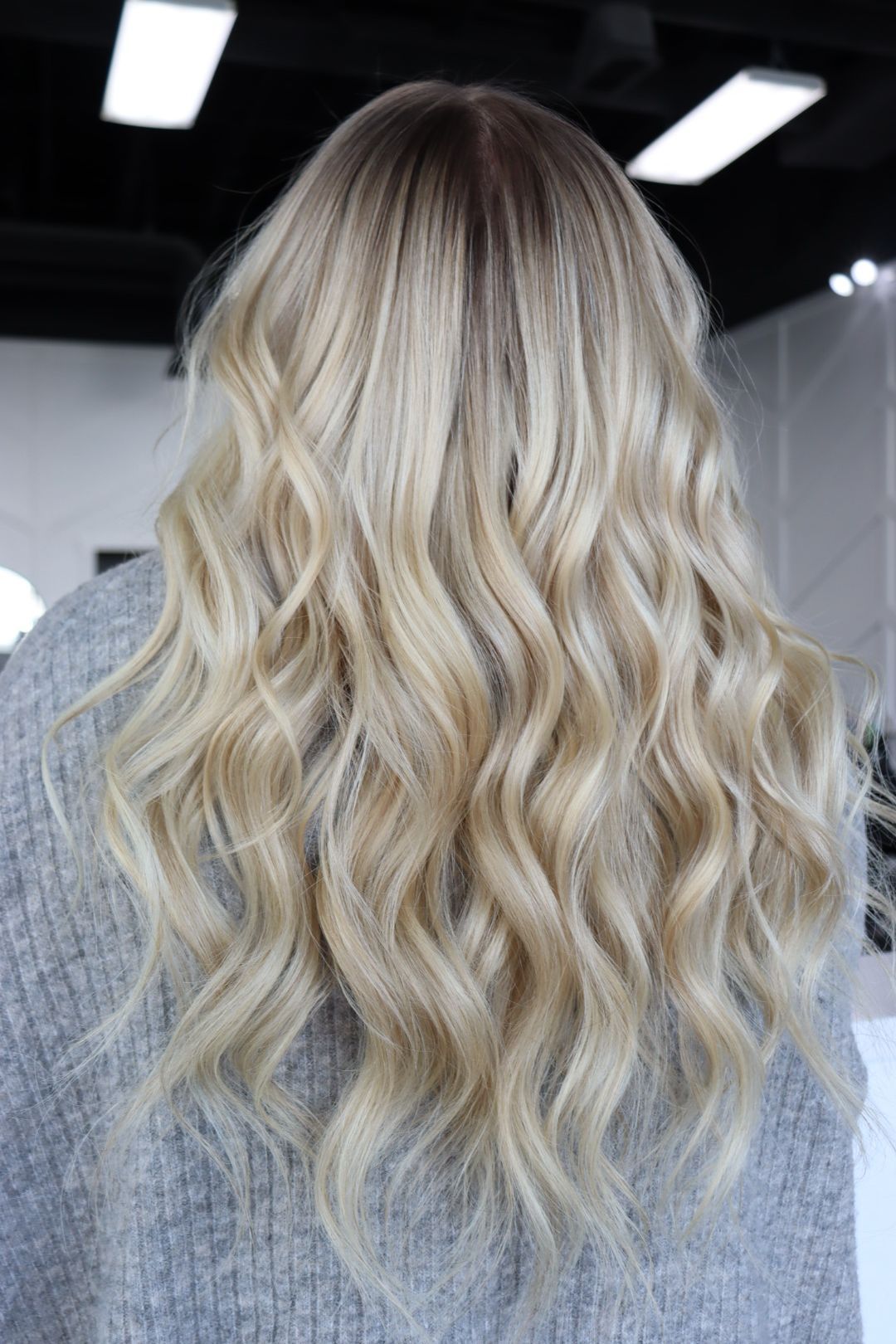 A woman with long blonde wavy hair is wearing a grey sweater.