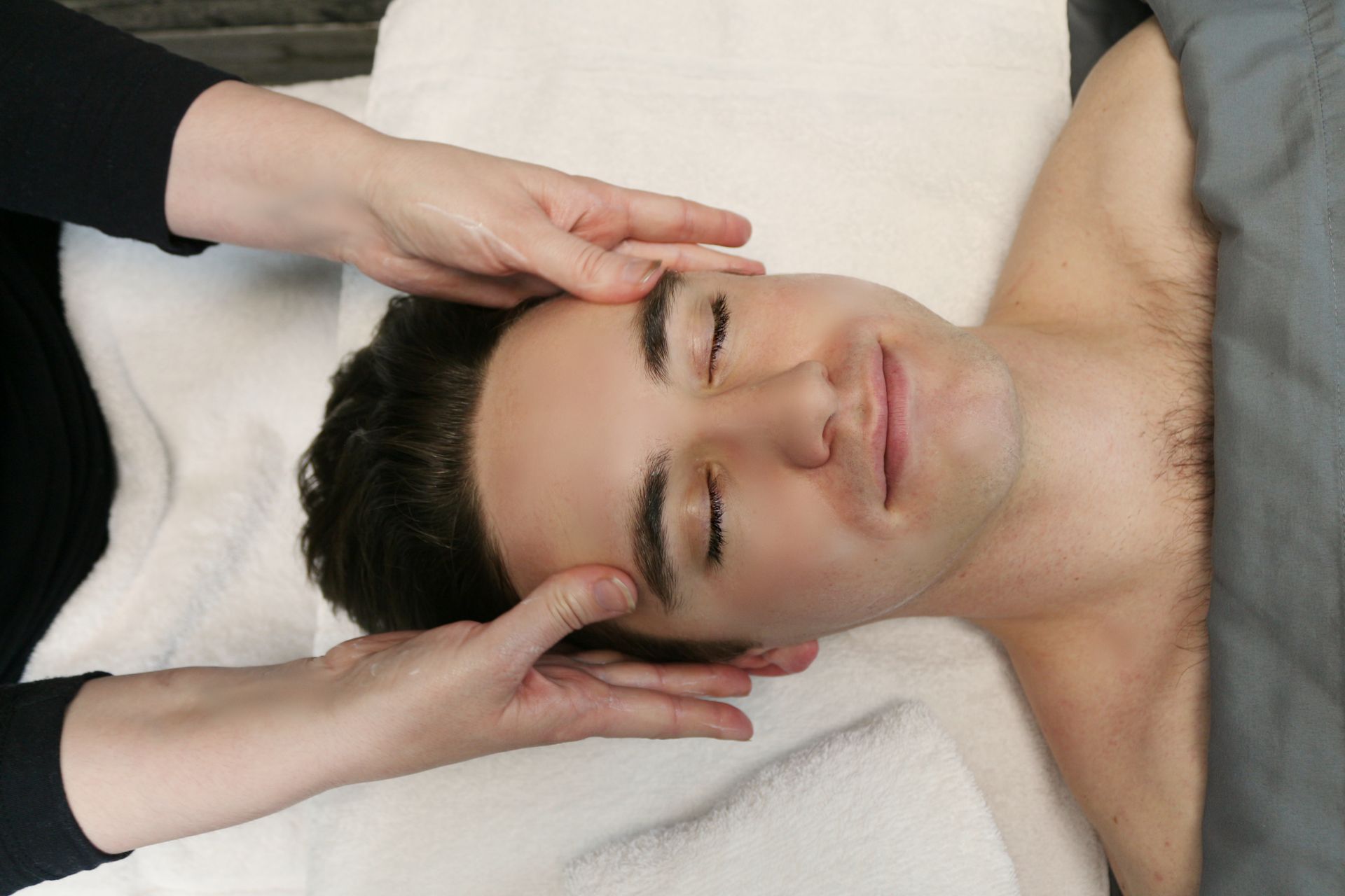 Man receiving a head massage with eyes closed; hands on forehead; white towel.