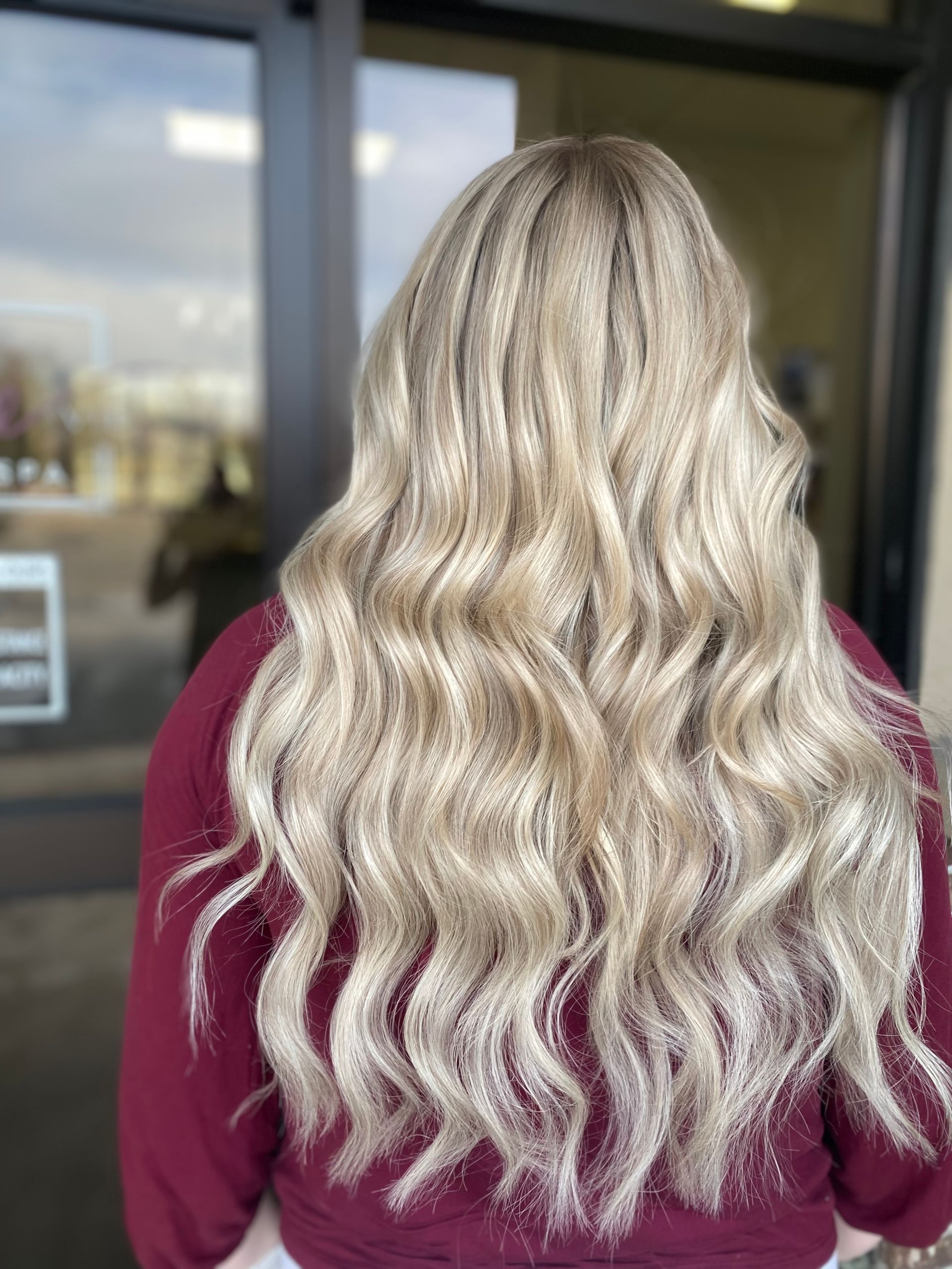 A woman with long blonde wavy hair is standing in front of a window.