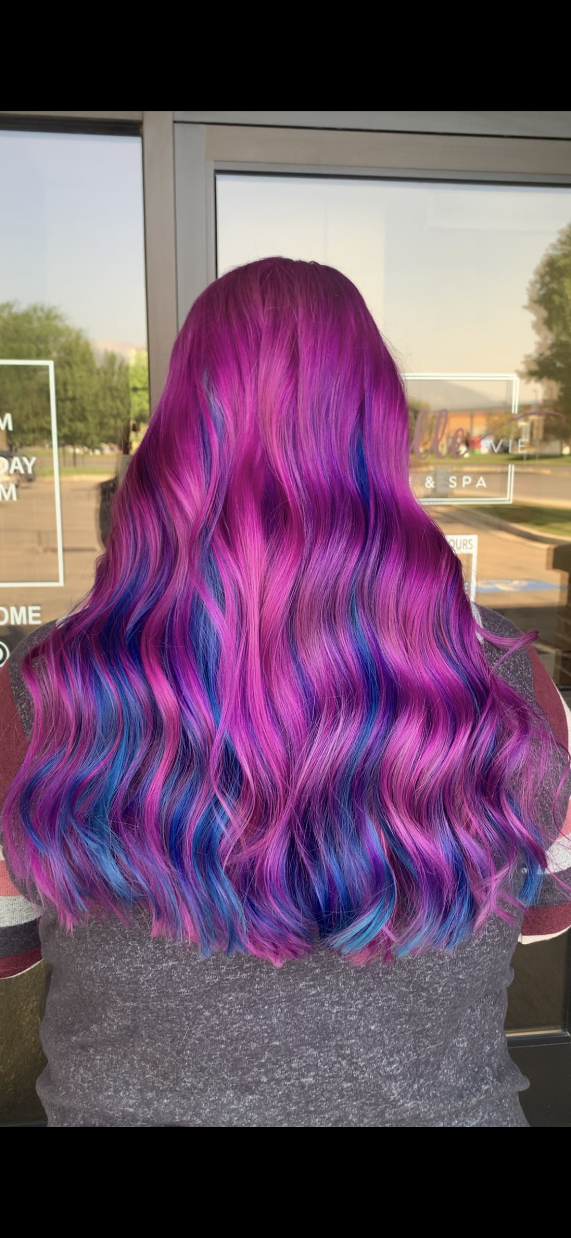 A woman with purple and blue hair is standing in front of a window.