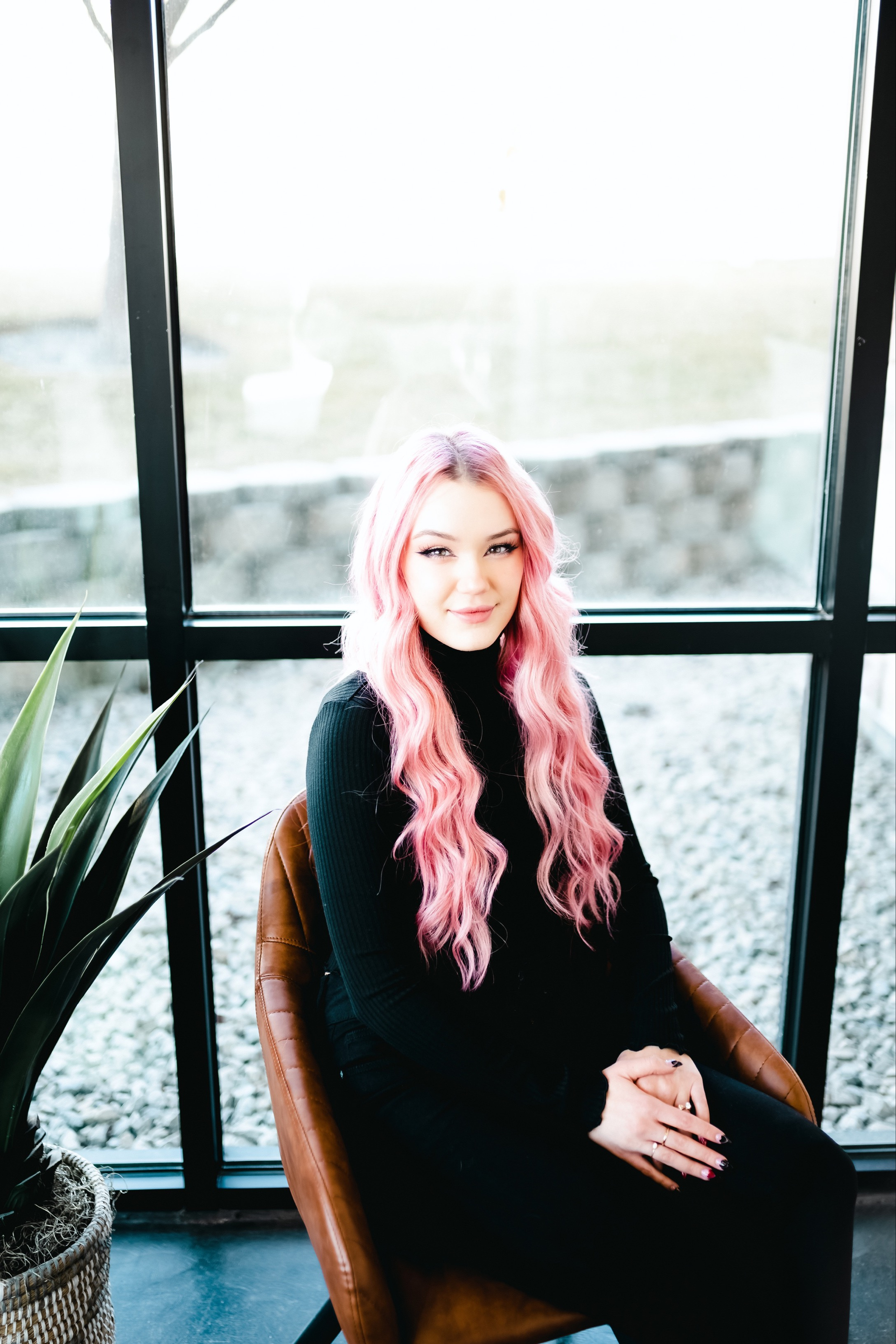 A woman with pink hair is sitting in a chair in front of a window.