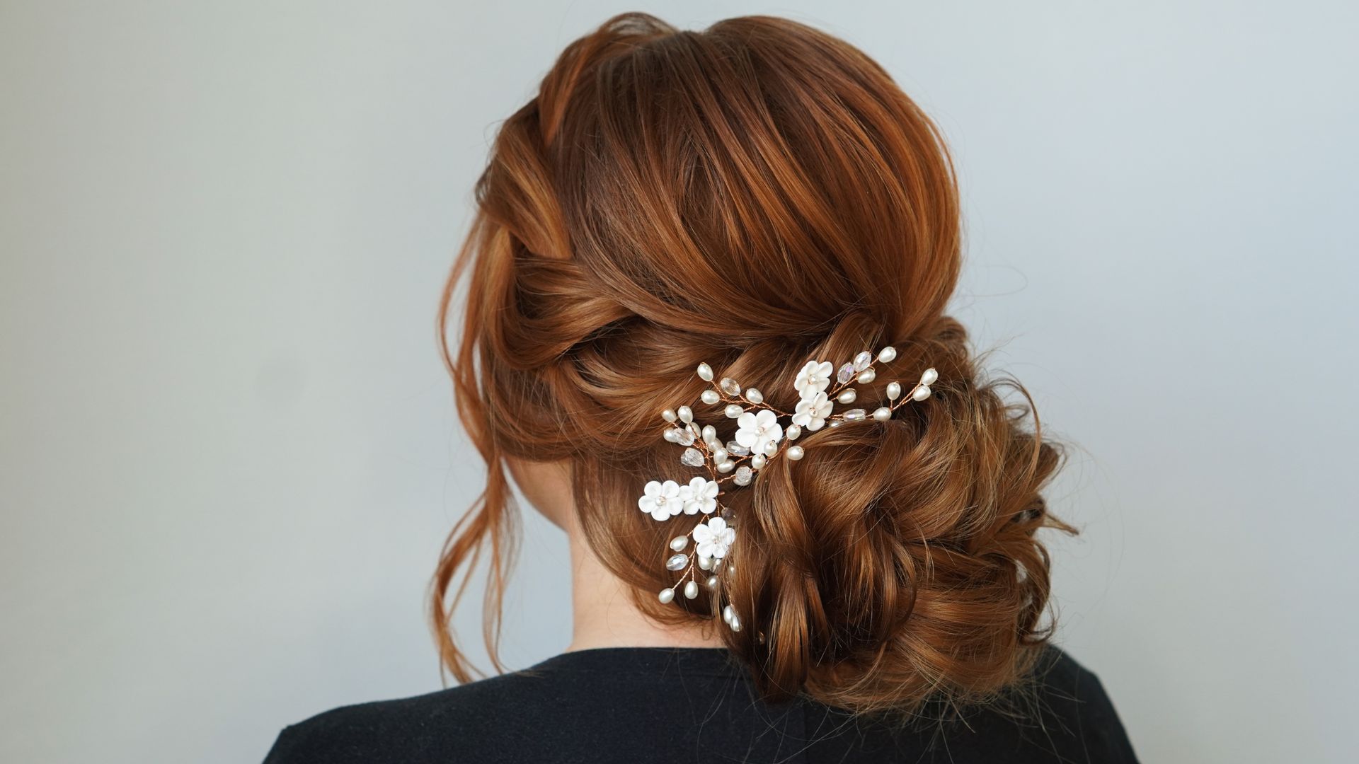 Woman with auburn hair in an elegant updo, adorned with floral hair accessory against a light grey background.