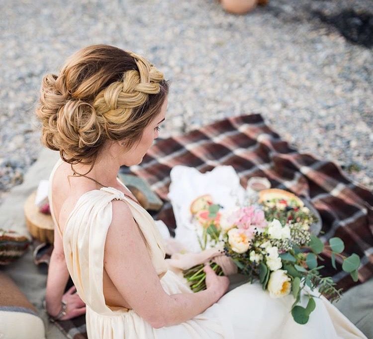 Woman with braided updo, wearing a cream dress, holding flowers, sitting on a picnic blanket.