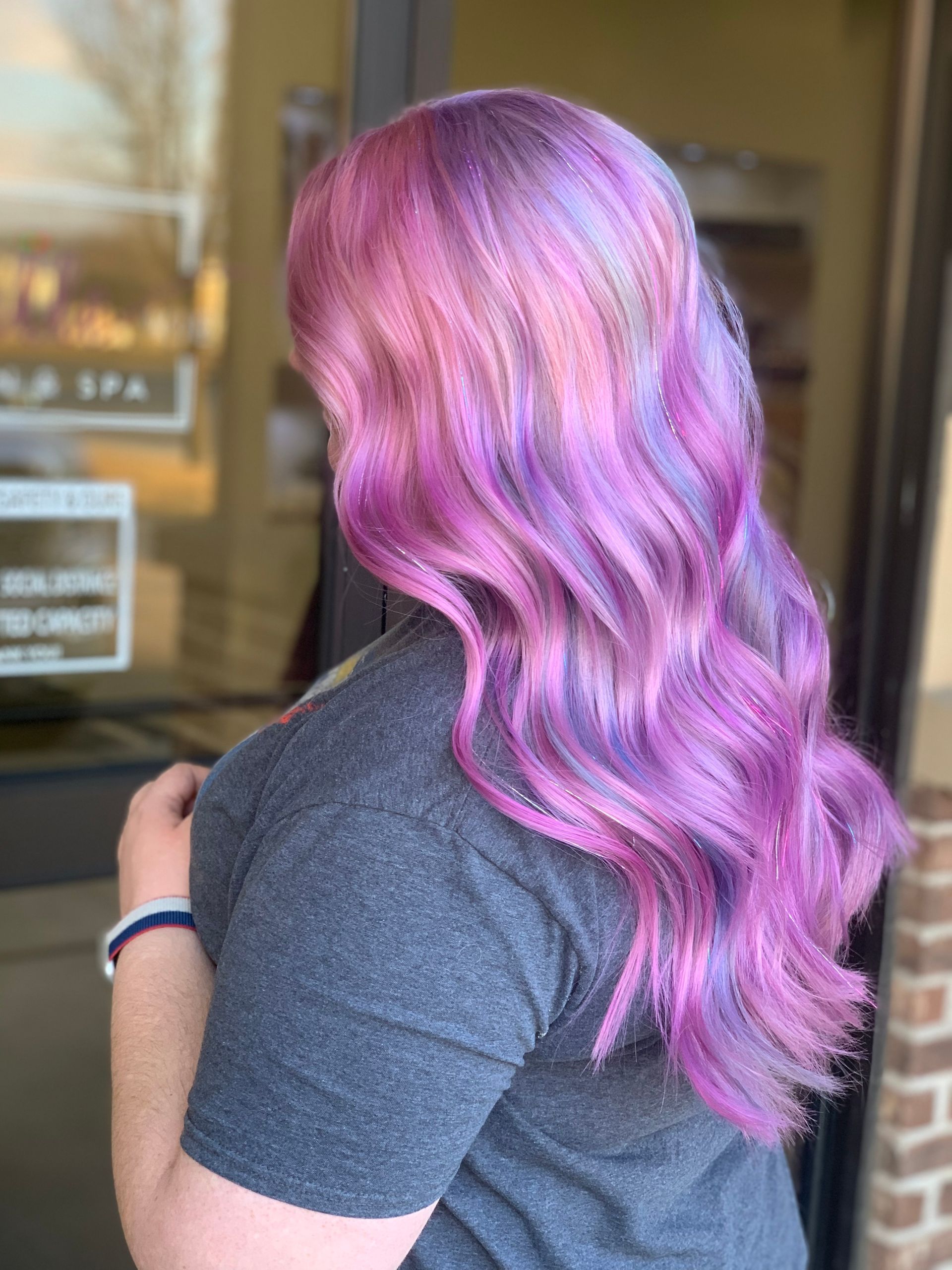 Woman with long, wavy, colorful hair, shades of pink, purple, and blue, in front of a spa.