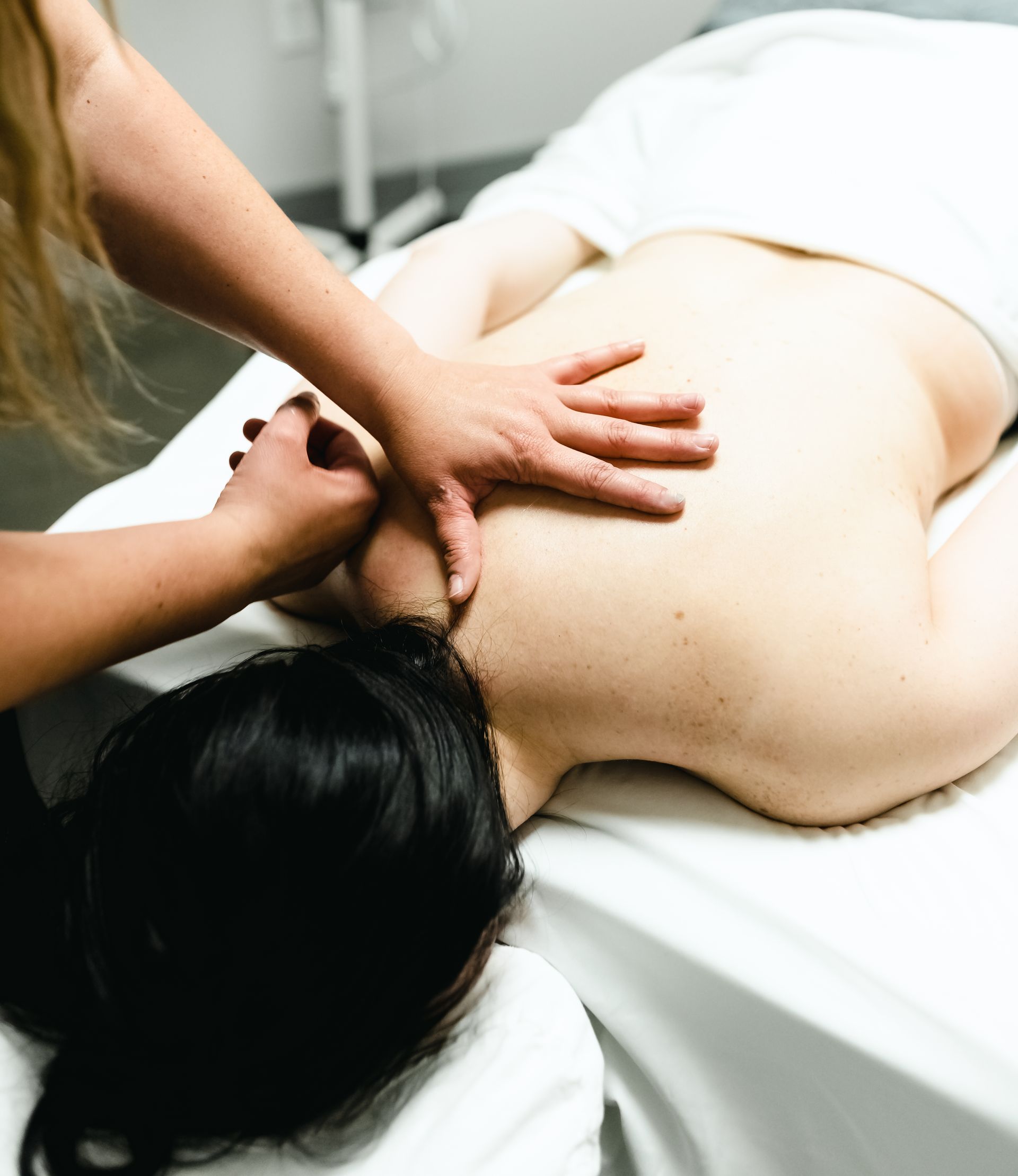 Person receiving a back massage; masseuse's hands on the client's back and shoulder, white sheets.