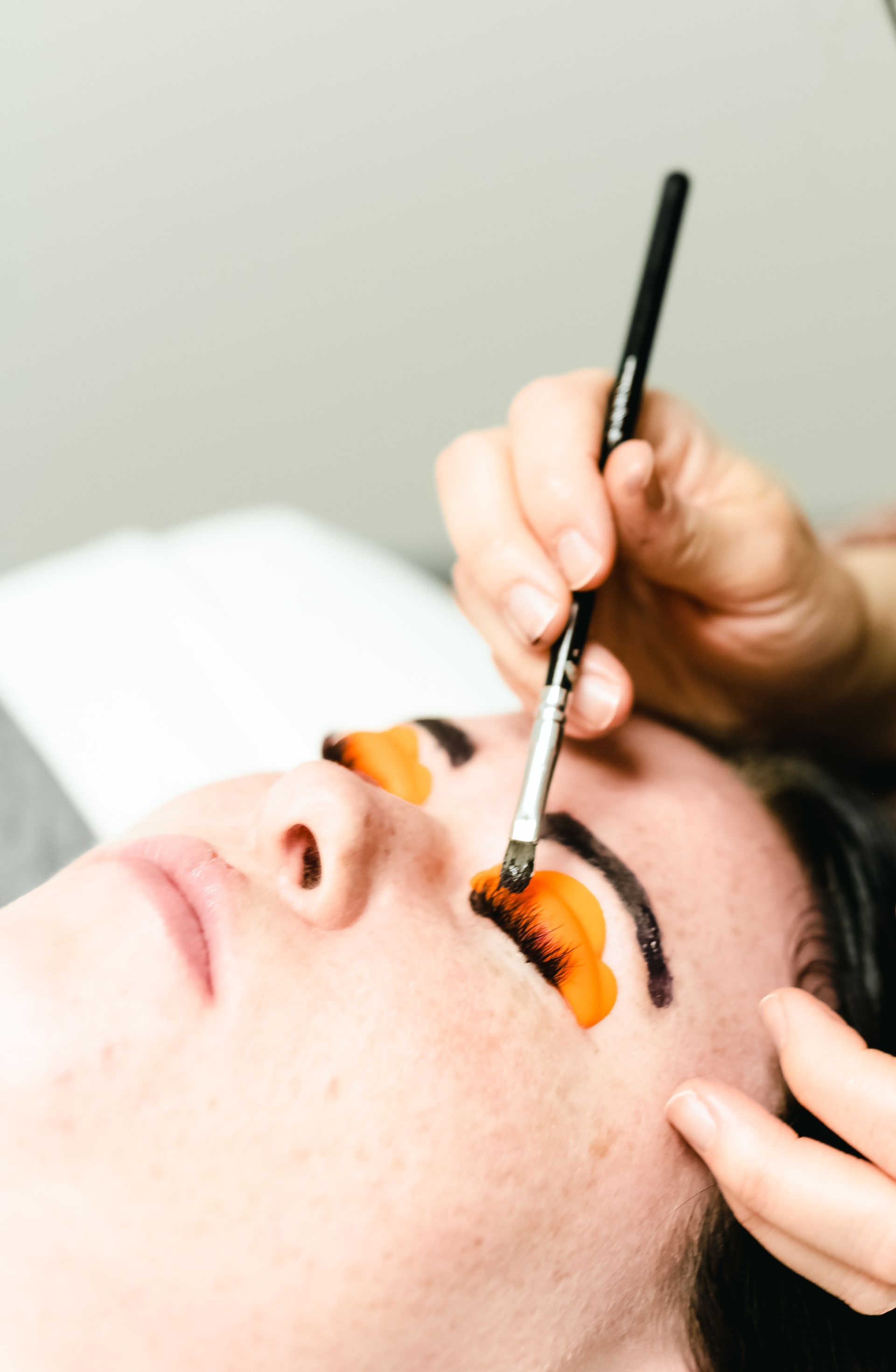 Person receiving an eyelash tint, with orange dye applied, being brushed by a professional.