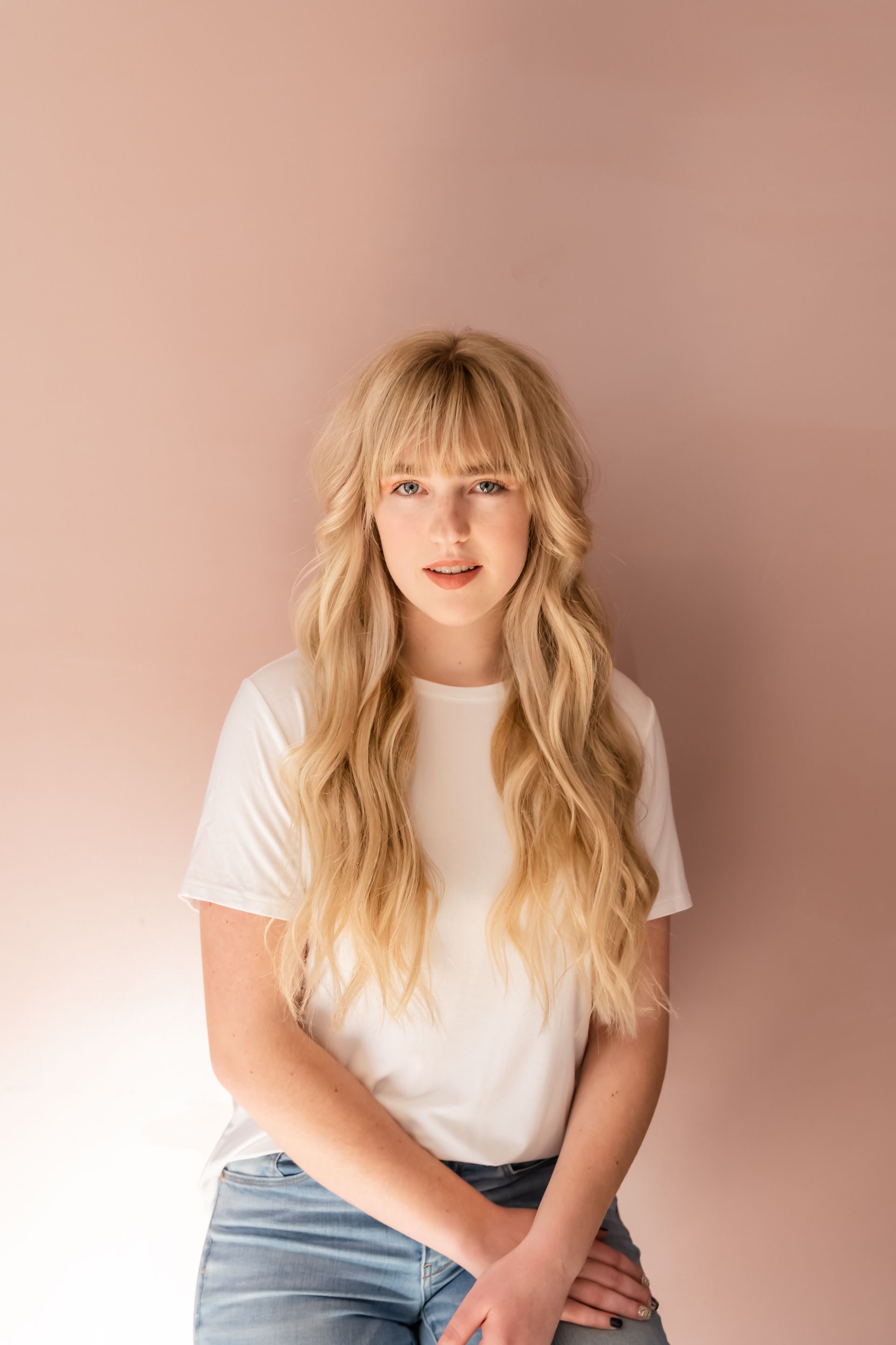 Blonde woman in white t-shirt and jeans leans against a pink wall.