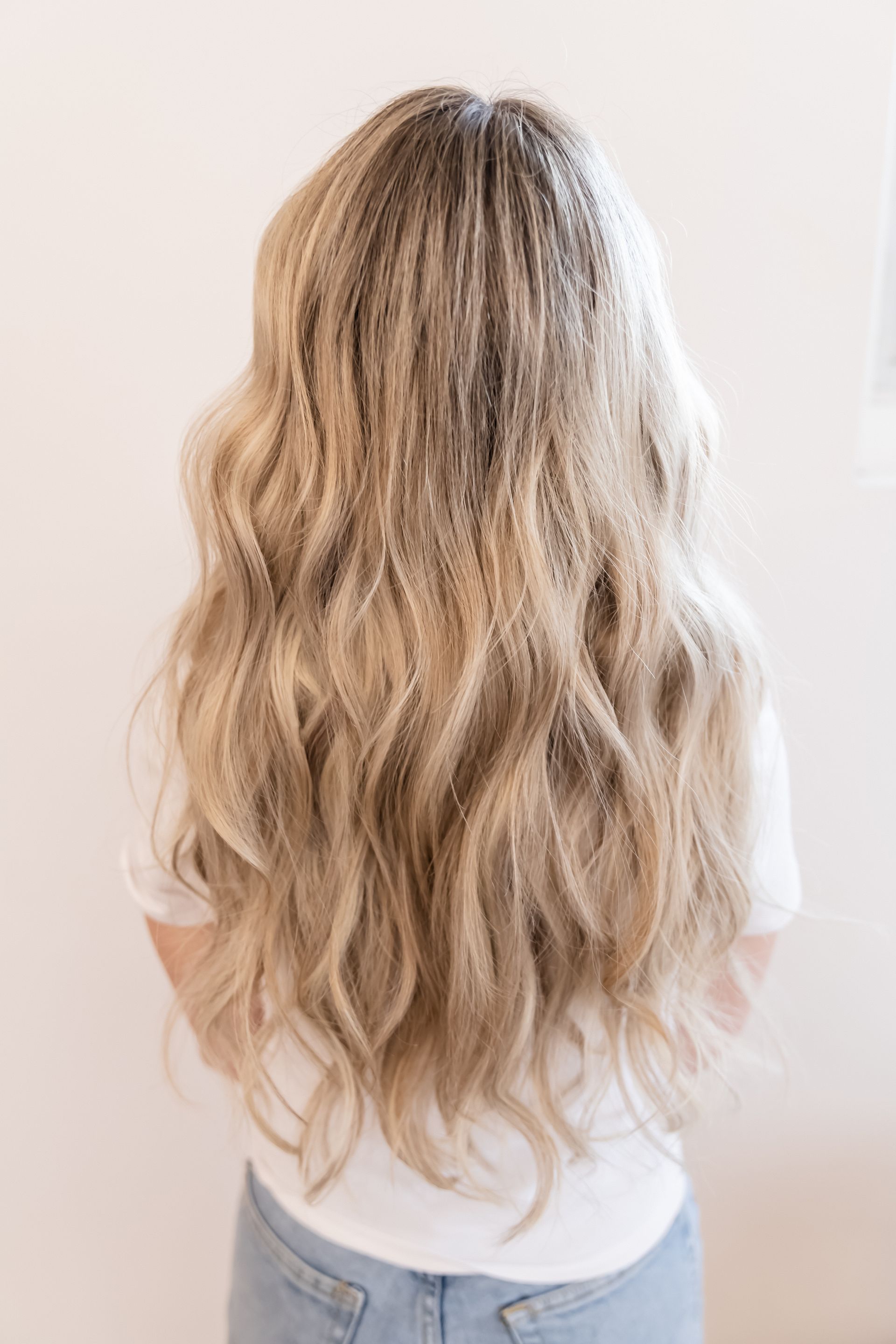 Blonde woman from the back, wearing a white shirt, with long wavy hair, standing against a neutral wall.