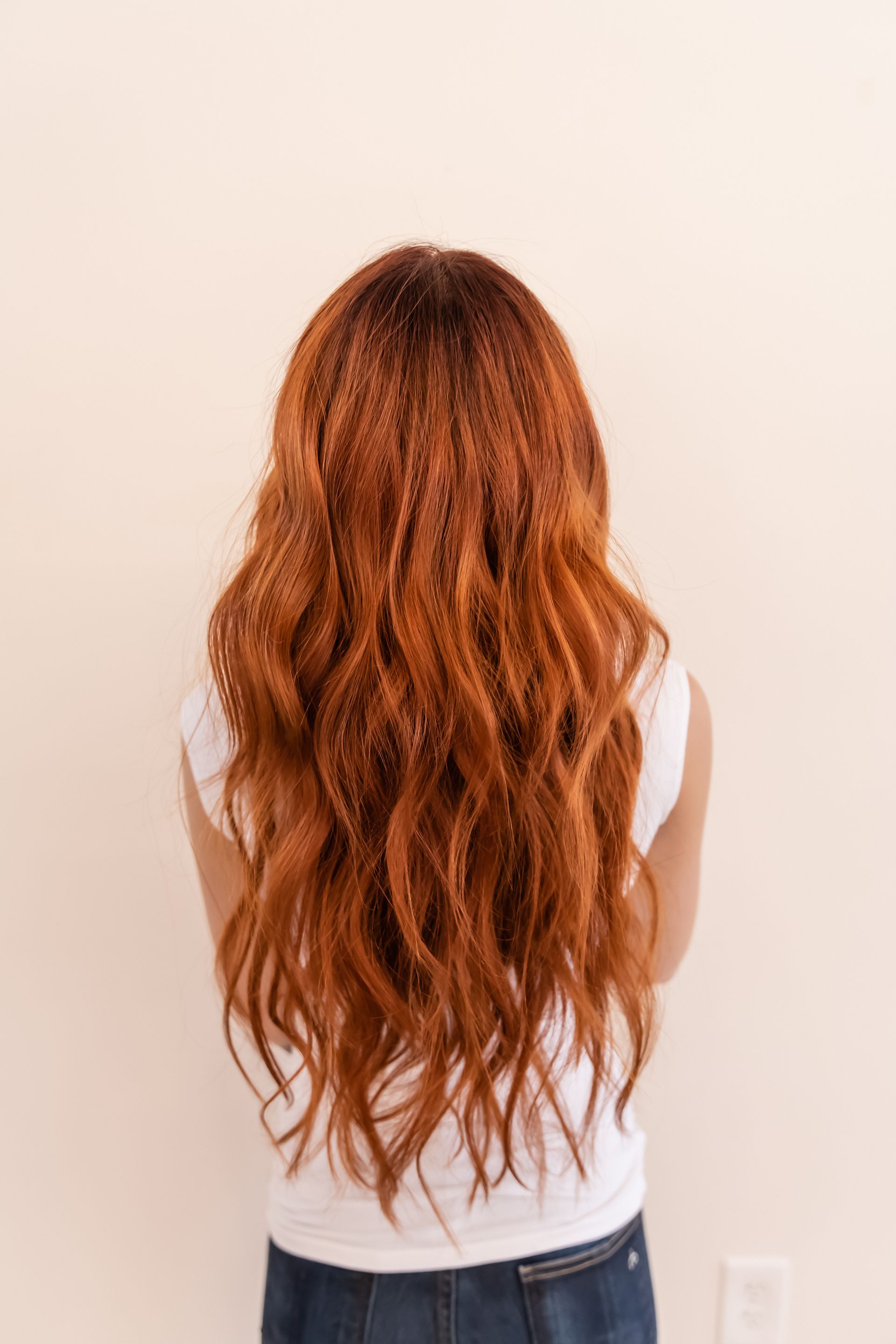 Person with long, wavy red hair wearing a white shirt and jeans, standing in front of a beige wall.