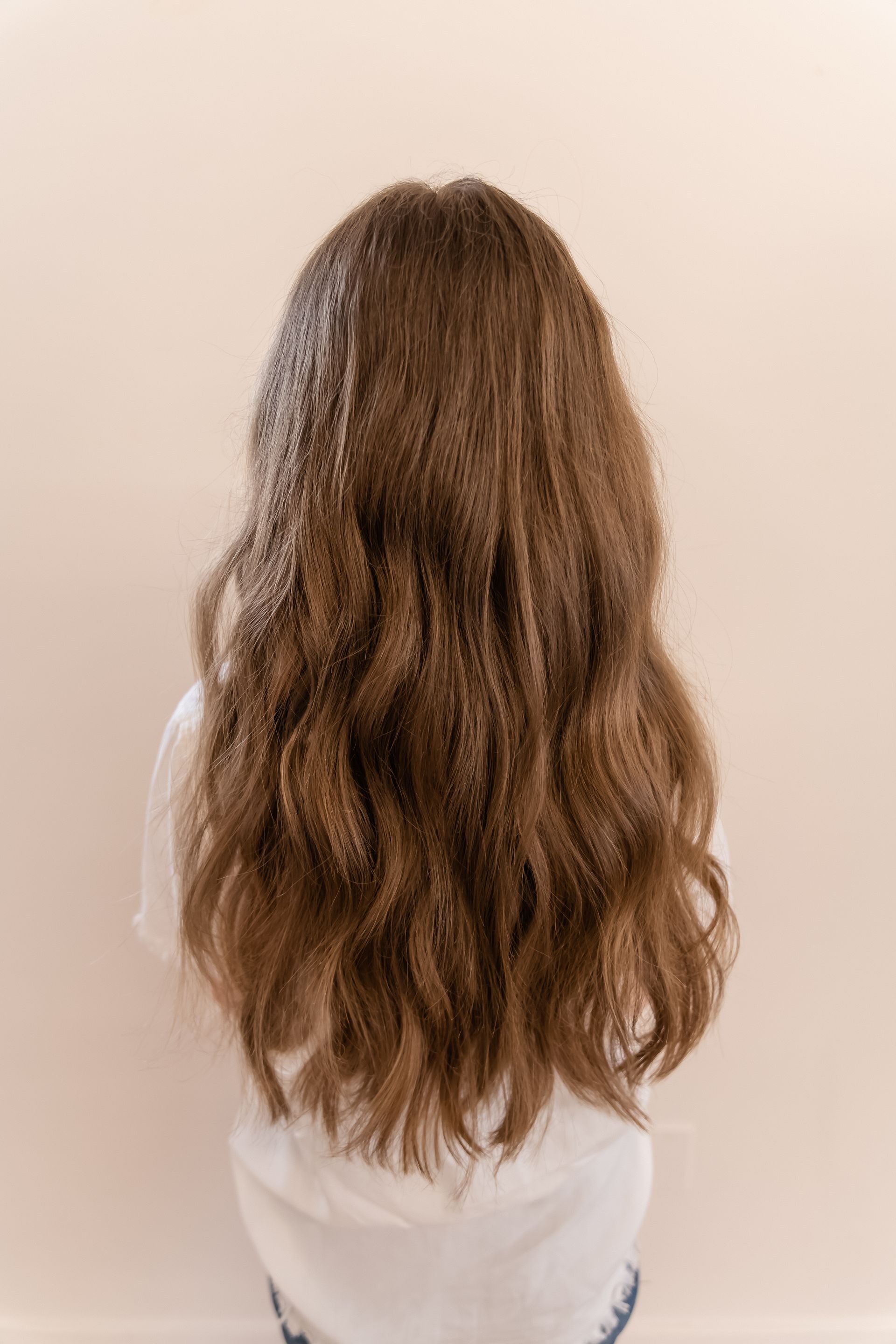 Person from the back with long brown wavy hair, wearing white top, against a light beige wall.