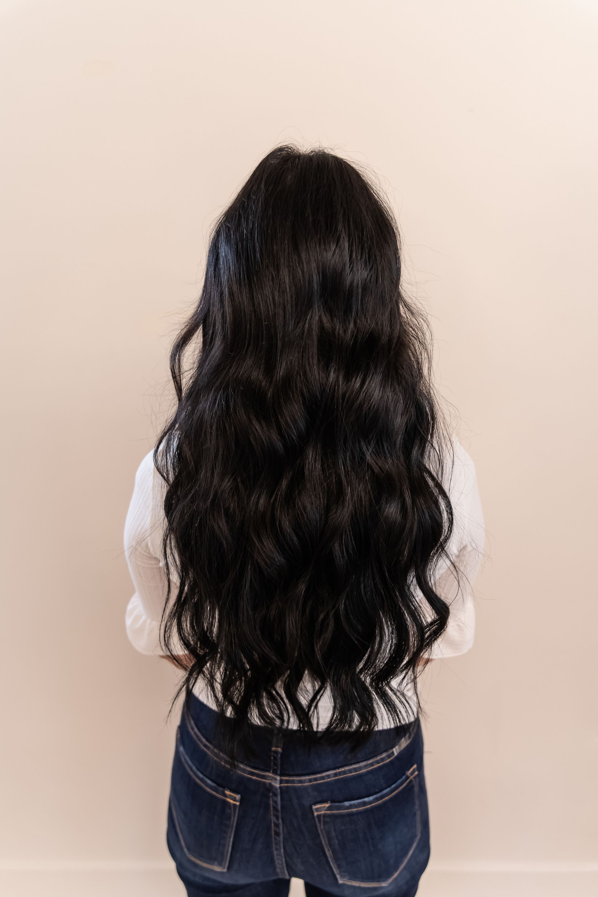 Person with long, dark curly hair wearing a white top and blue jeans, facing a plain wall.