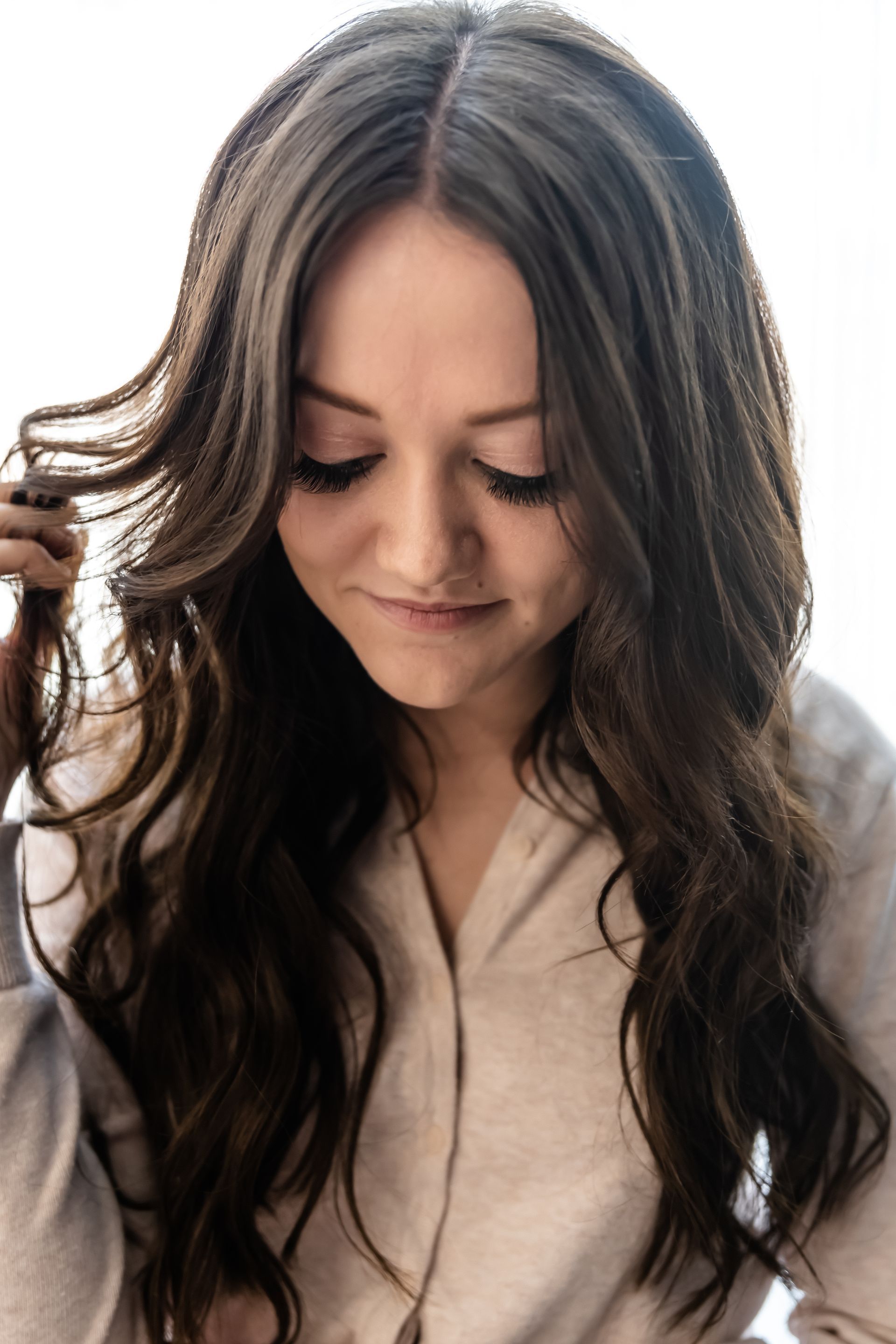 Woman with long brown hair, looking down, wearing a light-colored button-up shirt, touching her hair.