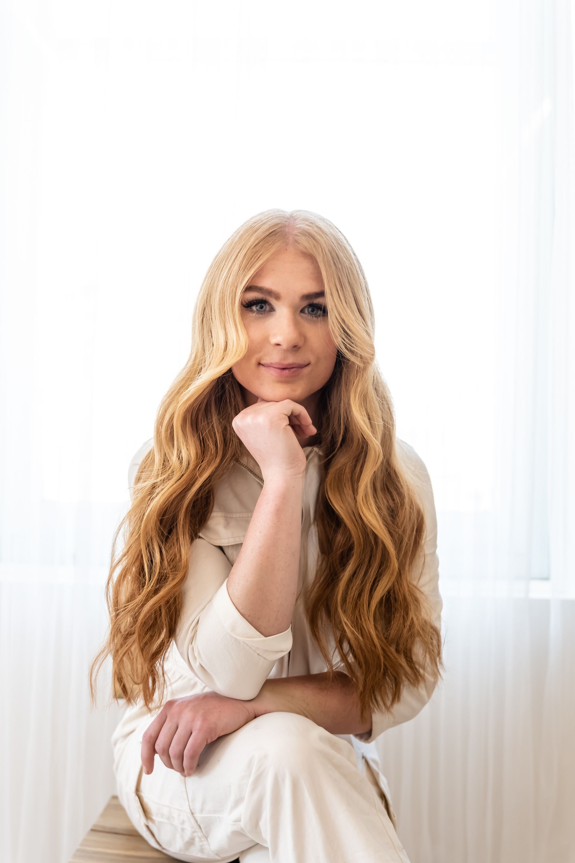 A woman with long blonde hair is sitting on a chair with her hand on her chin.