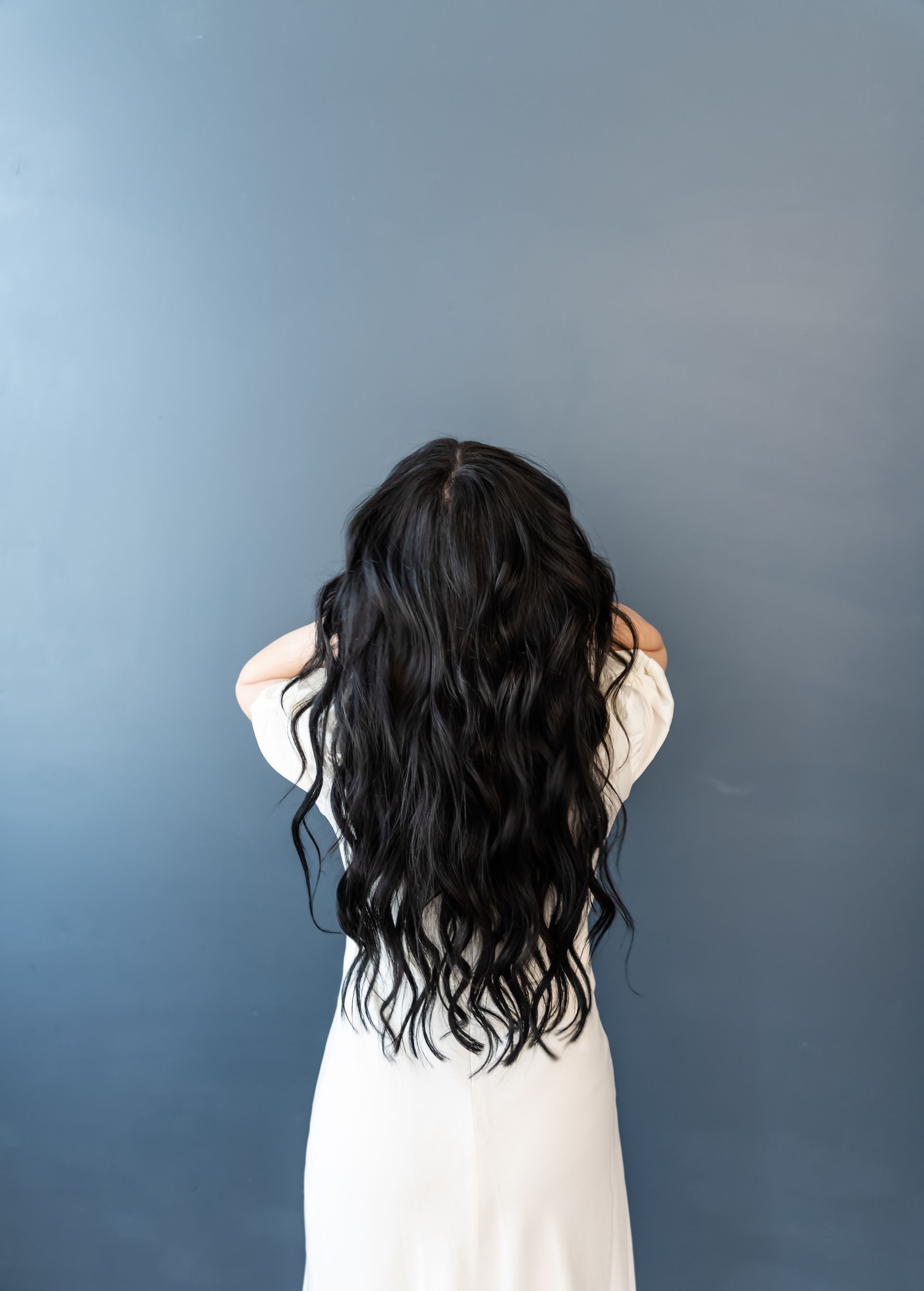 Woman with long black wavy hair and white dress, back to camera, against a blue wall.