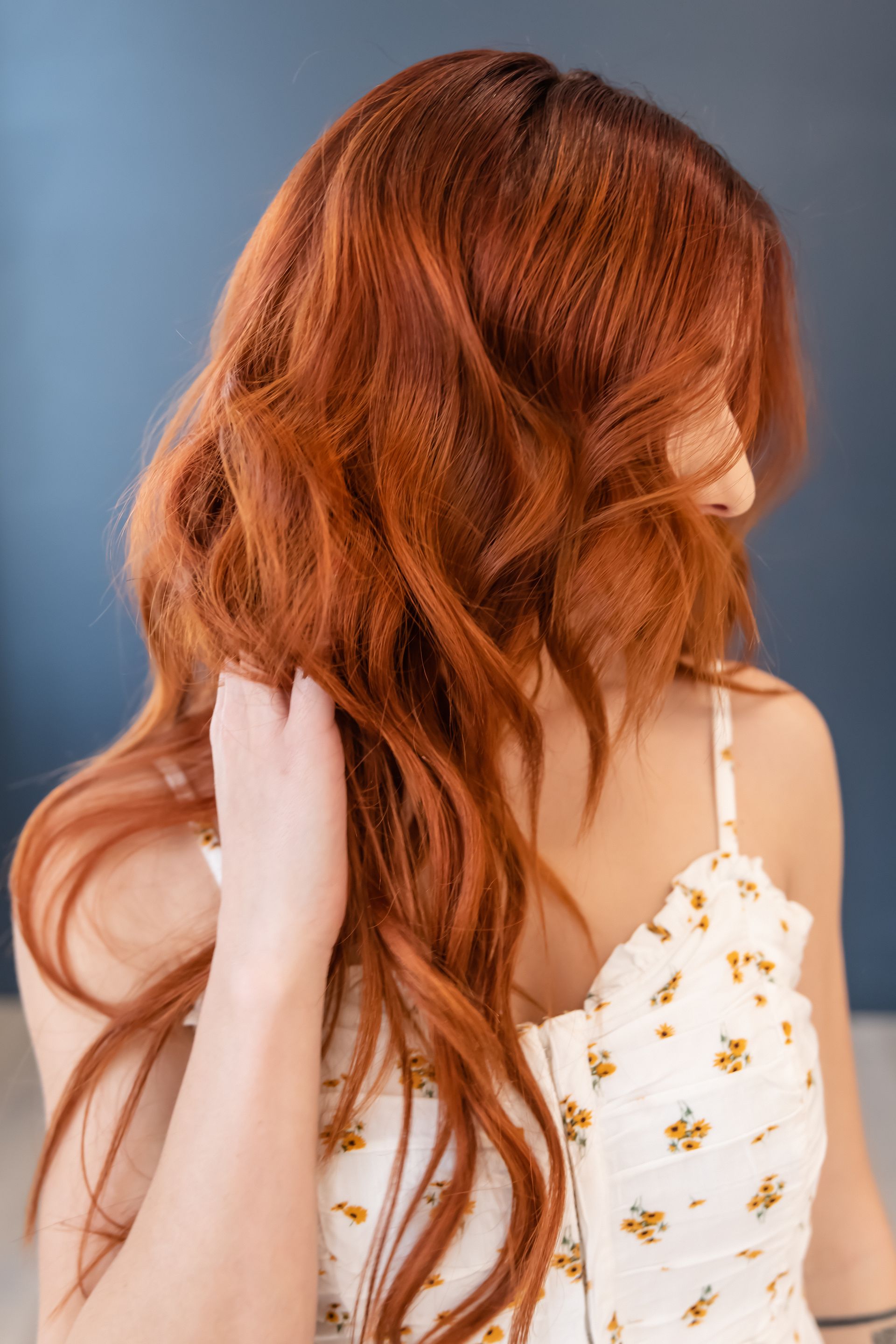 Woman with red hair, wearing a floral dress, touching her hair; against a dark blue background.