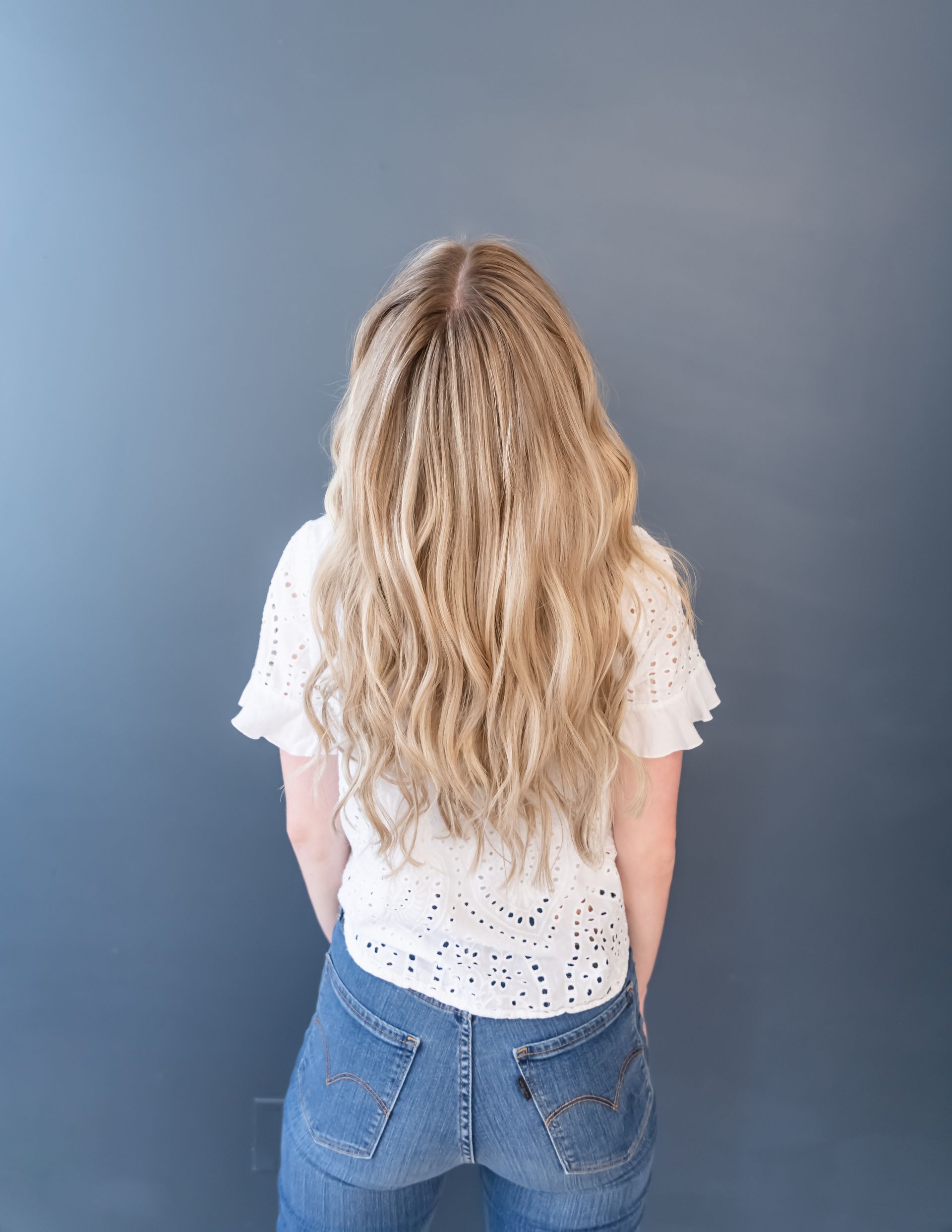 Woman with long, crimped blonde hair, wearing a white eyelet shirt and jeans, facing a gray wall.