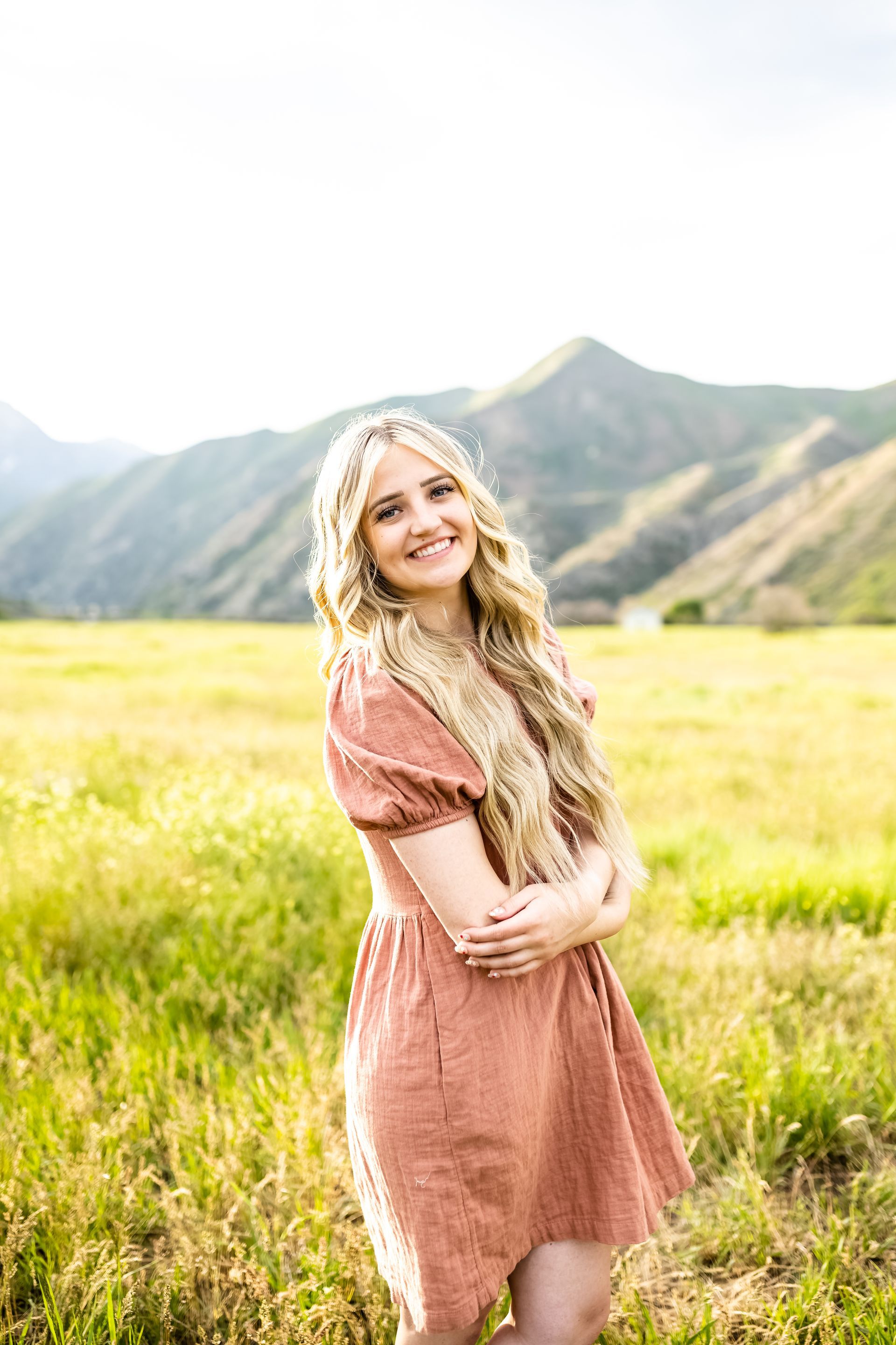 A woman in a pink dress is standing in a field with mountains in the background.