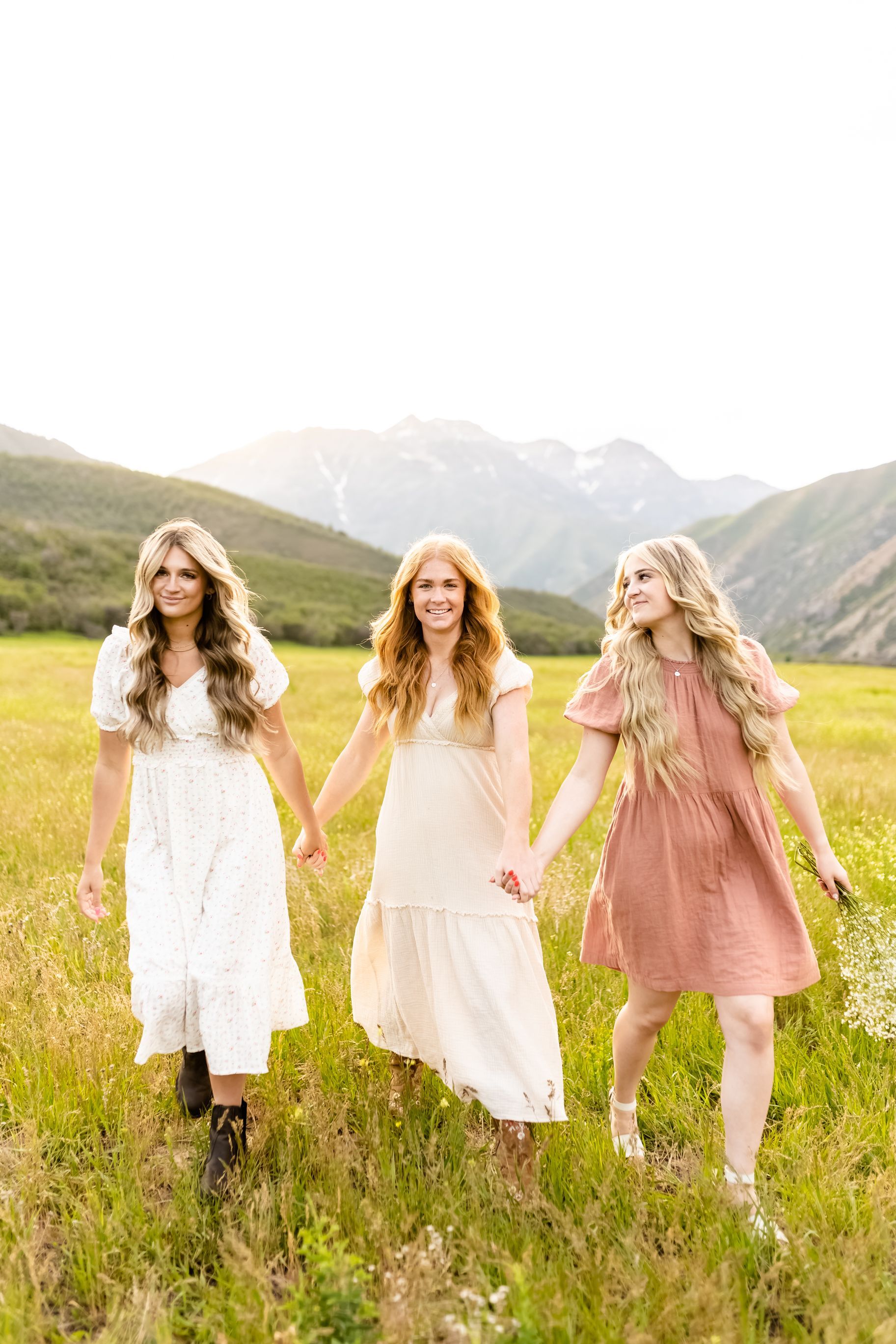 Three women in dresses are walking through a field holding hands.
