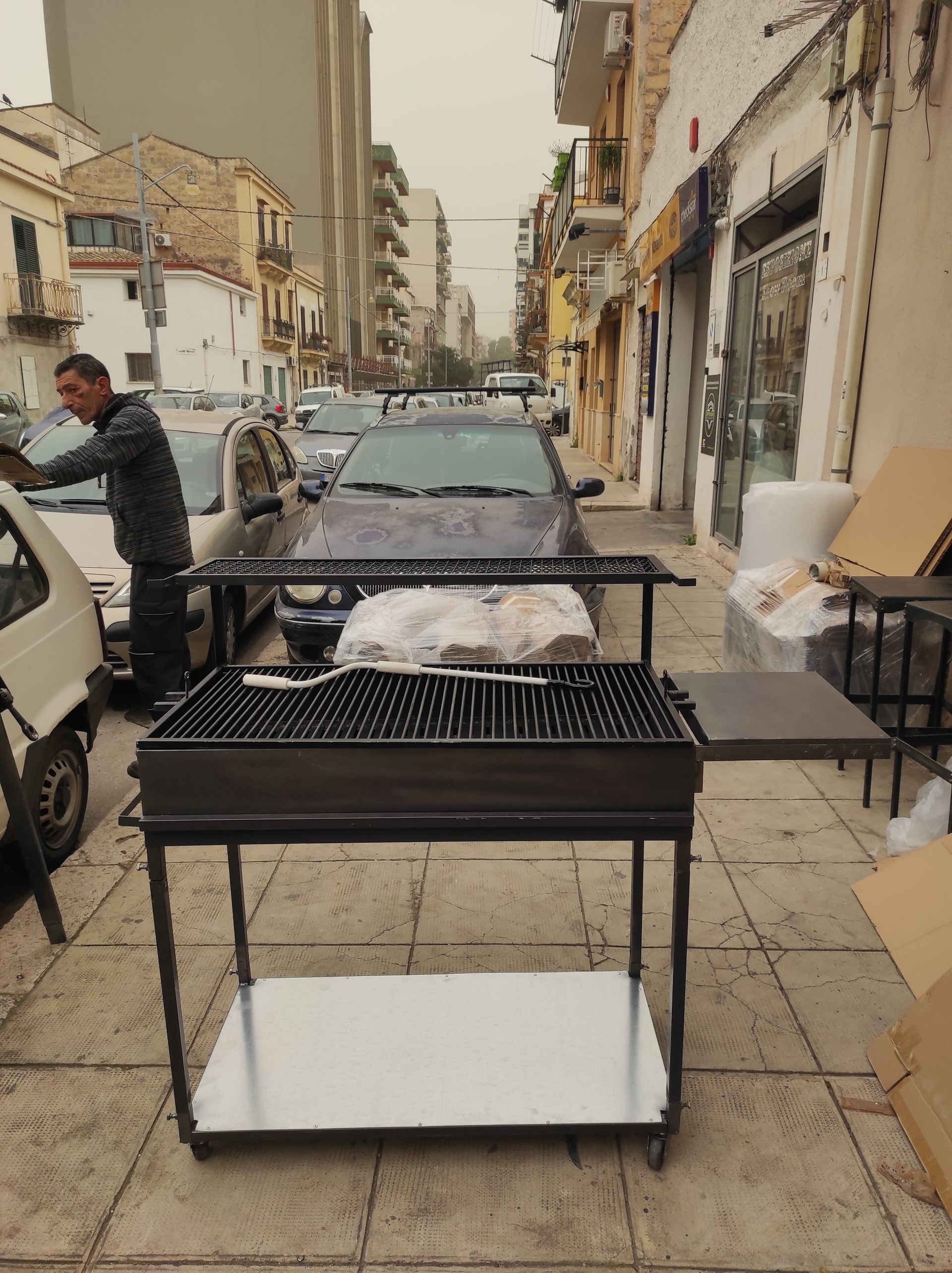 Uomo che griglia la carne su una griglia a carbone su un marciapiede accanto alle auto parcheggiate.