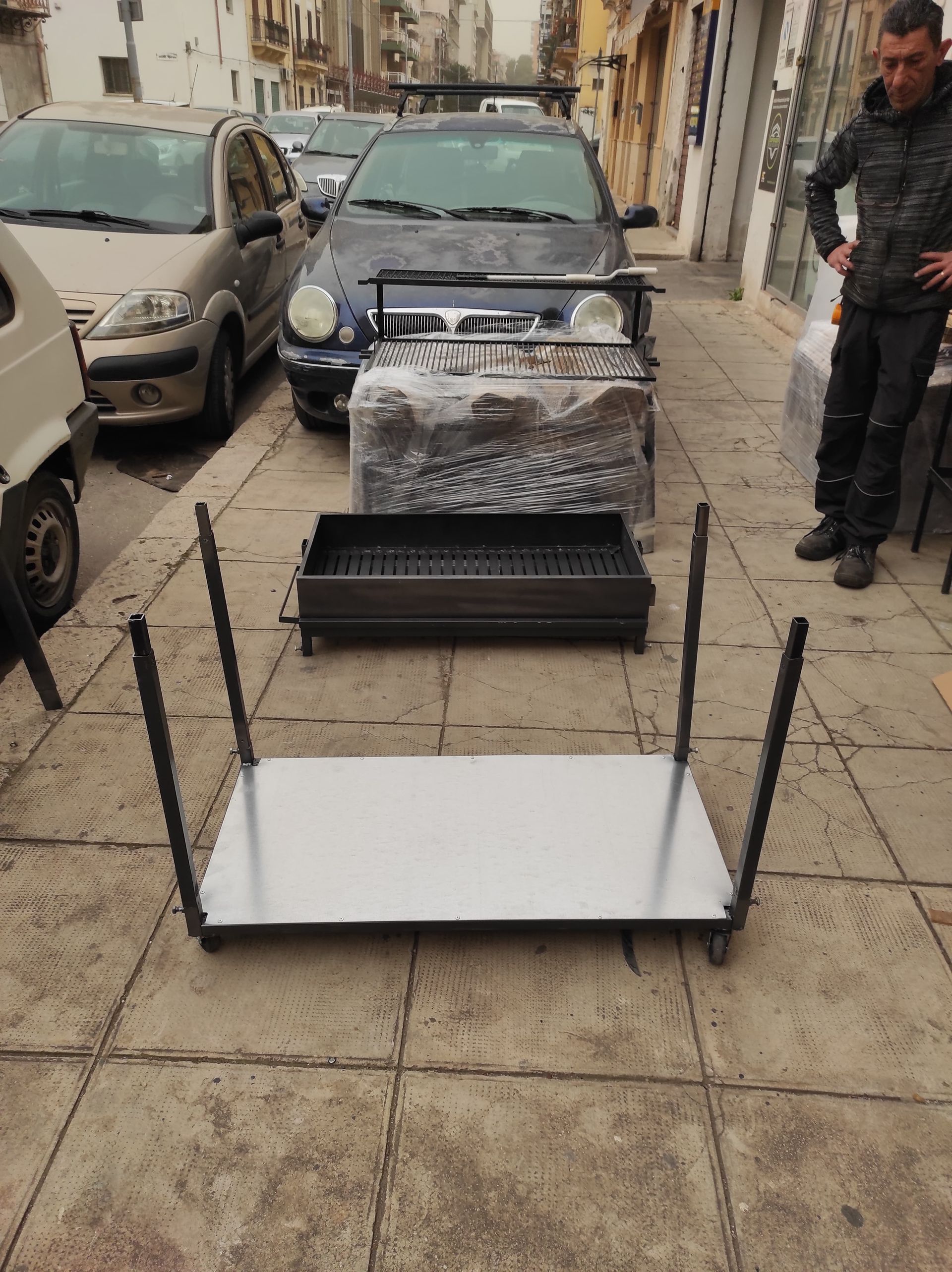 Una griglia di metallo, gambe e componenti sono posizionati su un marciapiede; un uomo è in piedi vicino alle auto parcheggiate.