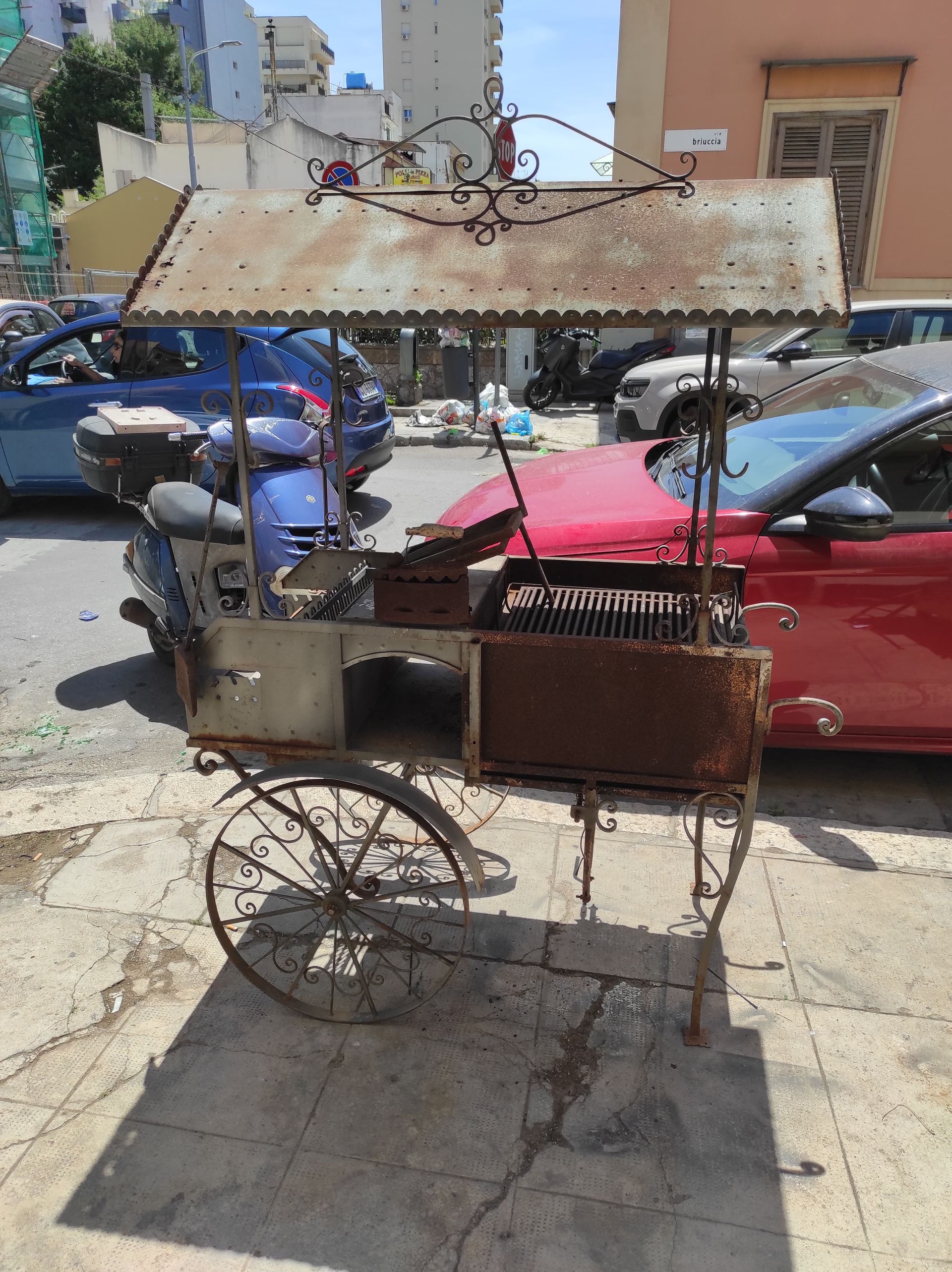 Un carretto arrugginito con un tendone che vende cibo da strada su un marciapiede, con delle auto sullo sfondo.