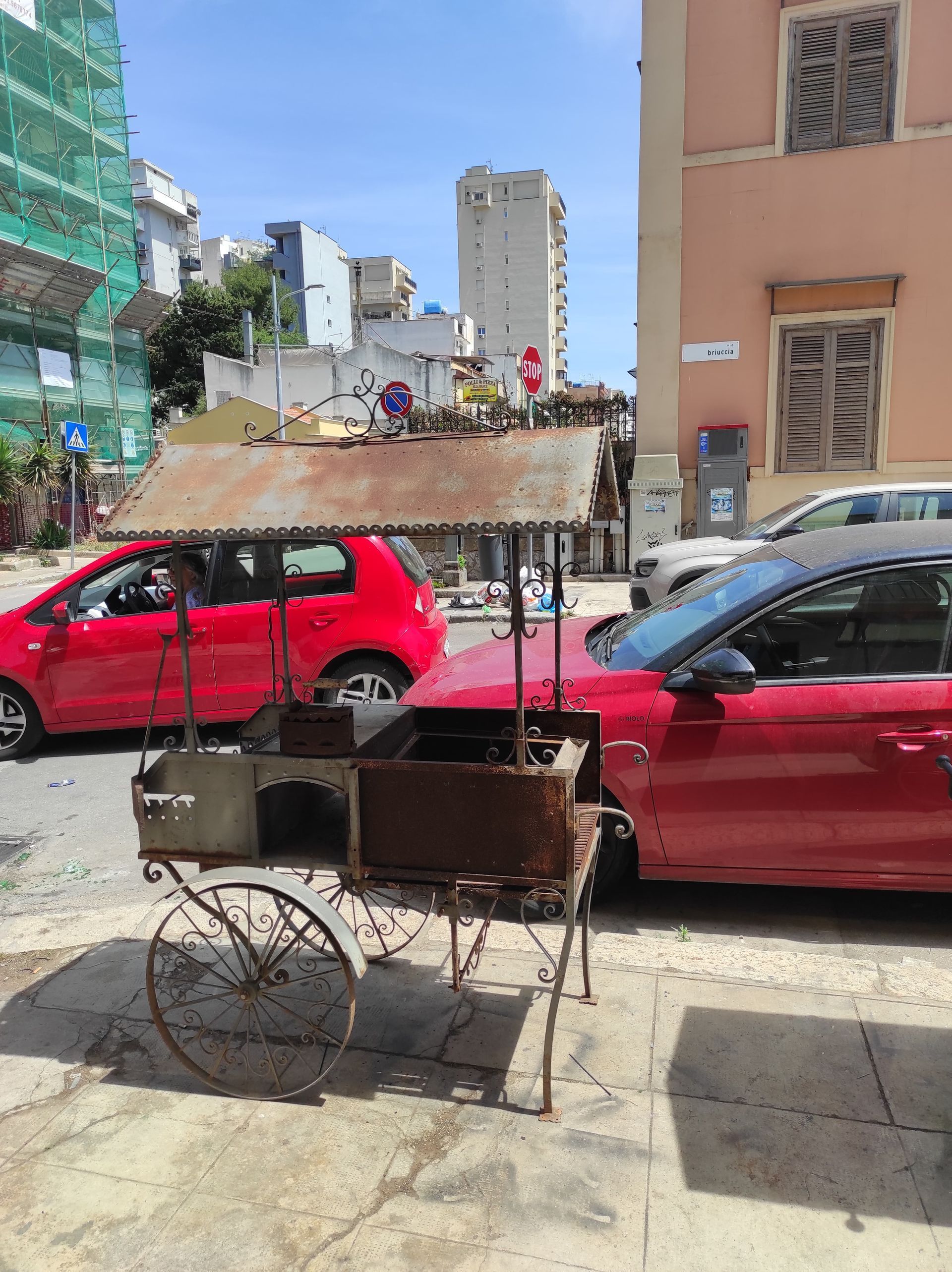 Vecchio carretto arrugginito di un venditore ambulante su un marciapiede con auto rosse ed edifici sullo sfondo in una giornata di sole.