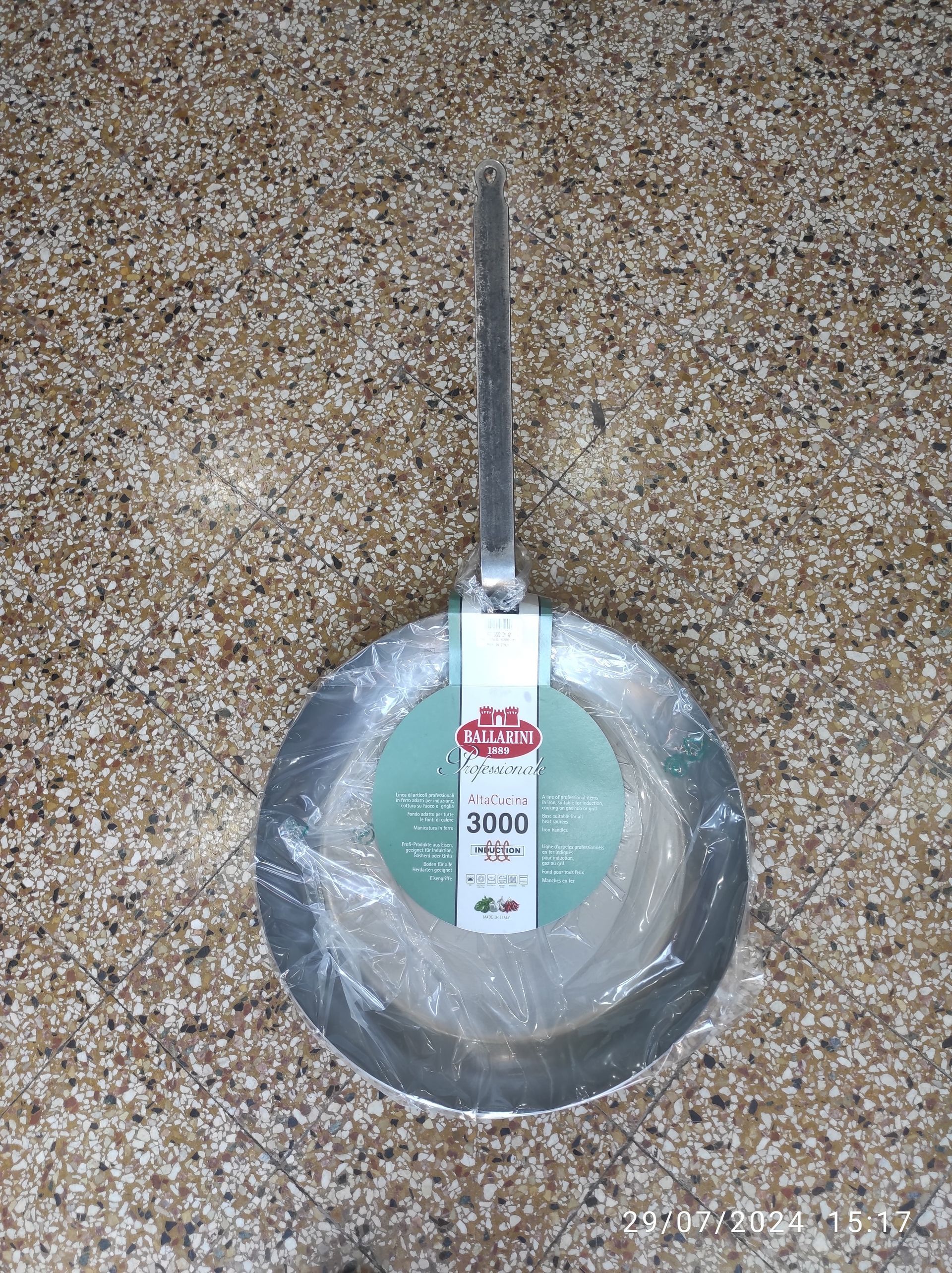 Un wok di metallo con un lungo manico, avvolto nella plastica, poggia su una superficie punteggiata.