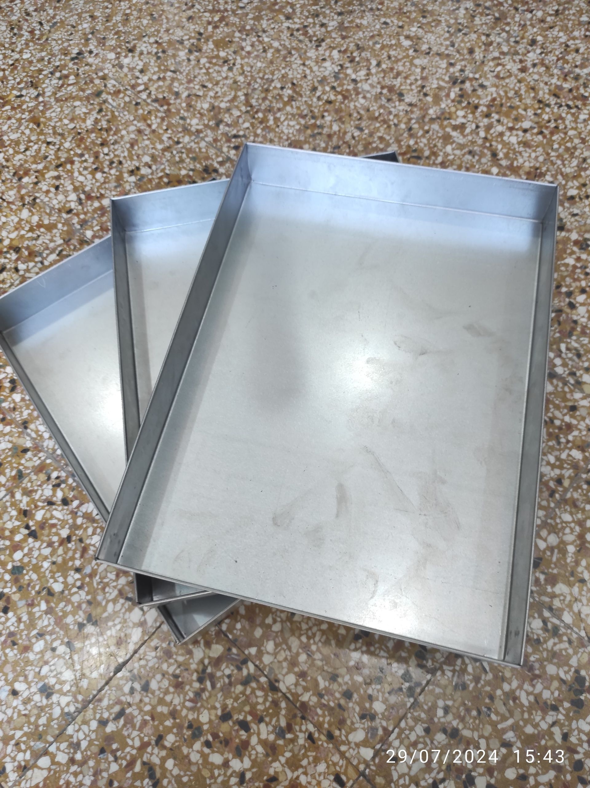 Tre vassoi rettangolari di metallo impilati su un pavimento punteggiato, probabilmente utilizzati per la cottura al forno o la preparazione del cibo.